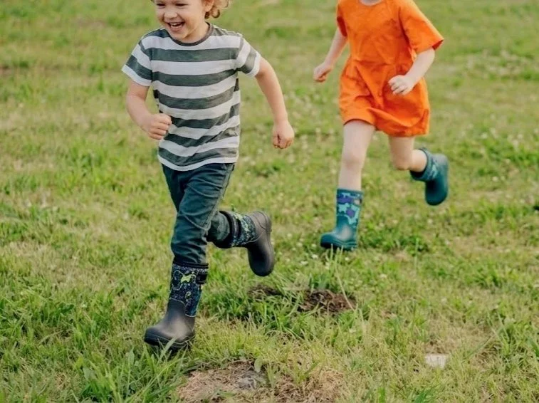 Two children running through a grassy field, wearing boots and casual clothes, enjoying outdoors.