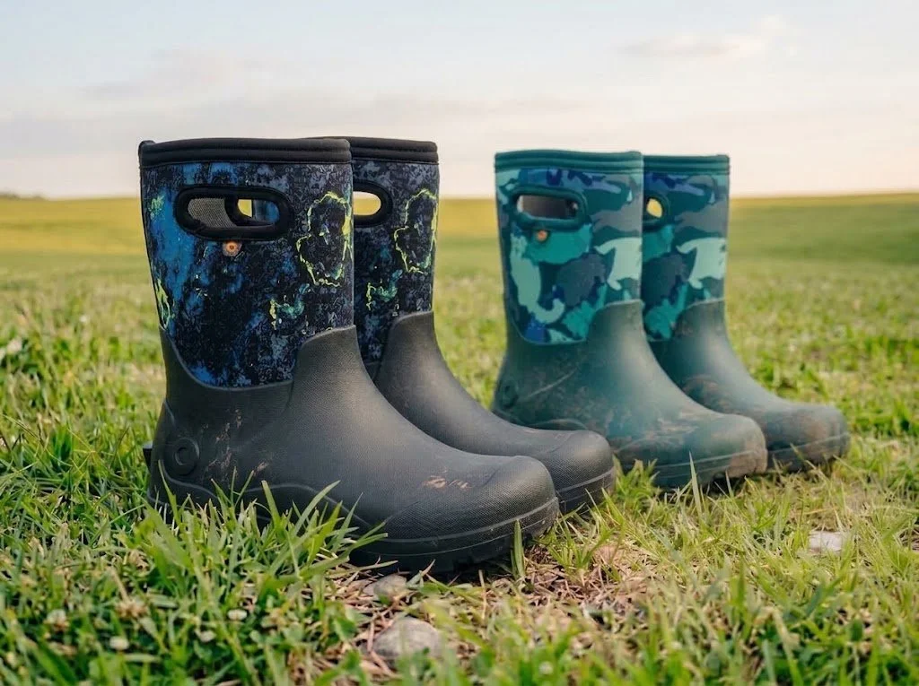 Two pairs of children's rain boots on grass, one with a dark blue and yellow pattern and the other with a blue camouflage pattern.