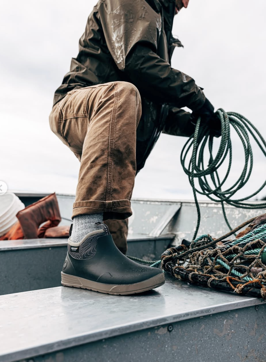 Person wearing brown pants and black waterproof boots on a boat, handling coiled ropes, with others and part of the boat visible under a cloudy sky.