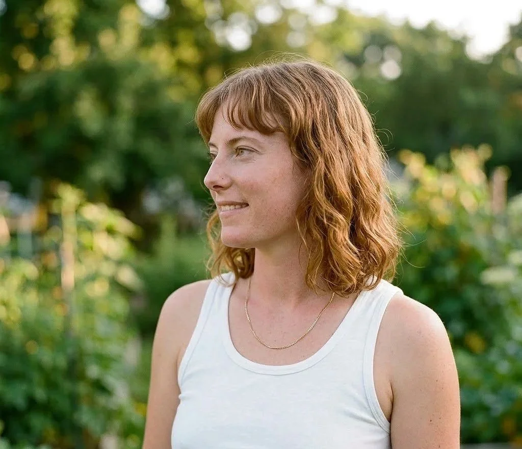 A woman with wavy, shoulder-length red hair smiling and looking to the side outdoors with green trees and sunlight in the background.