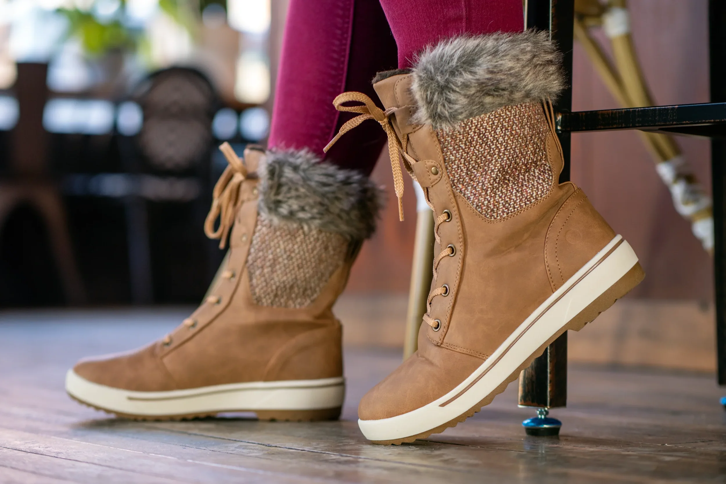 Pair of tan winter boots with faux fur trim and decorative fabric panels, placed on a wooden floor near a black metal chair with a small footrest. One boot has a small, blue, skull-shaped, studded accessory on the sole.
