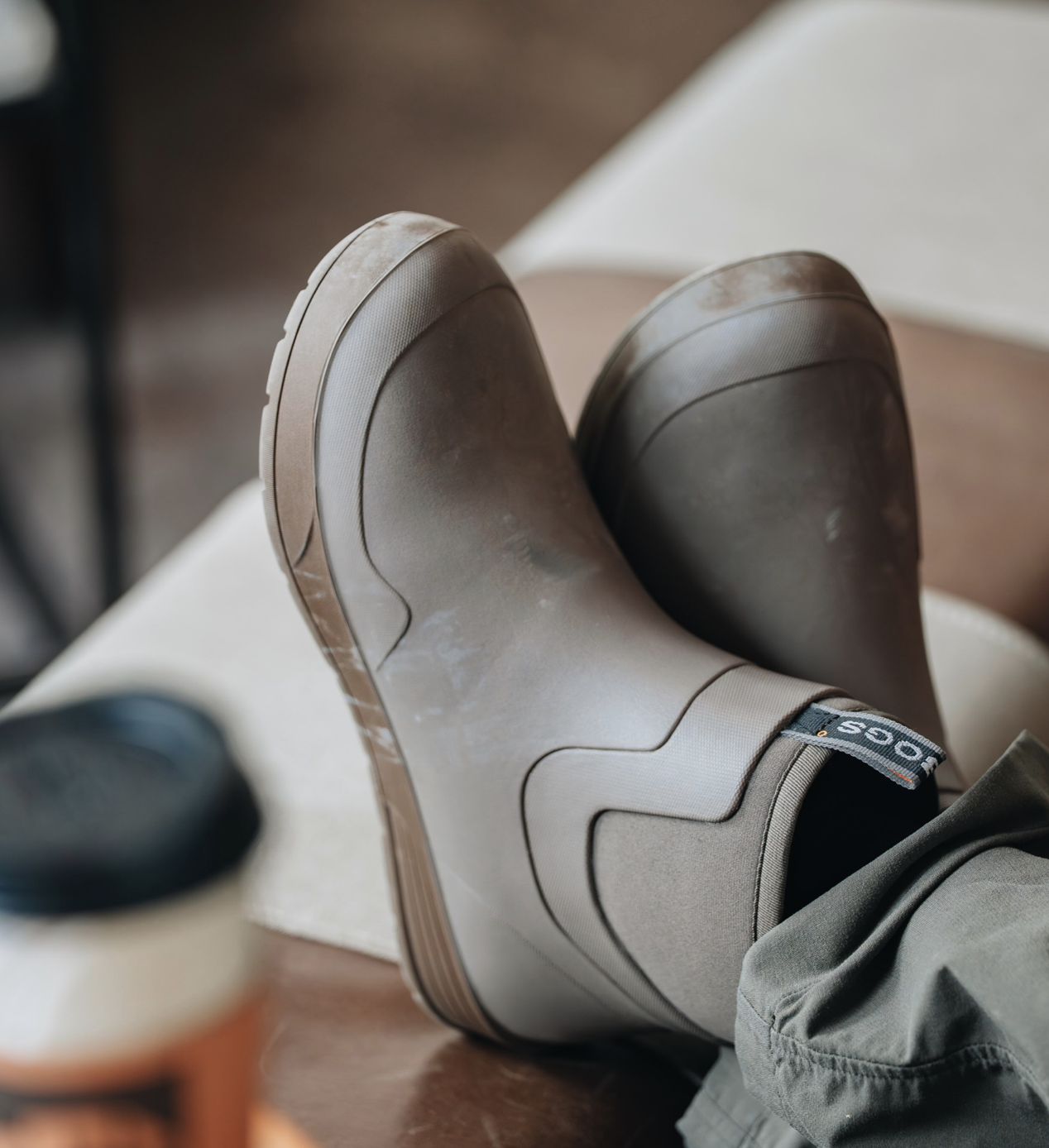 Person wearing beige rain boots with a brown sole, sitting on a white surface with legs crossed, in a casual indoor setting.