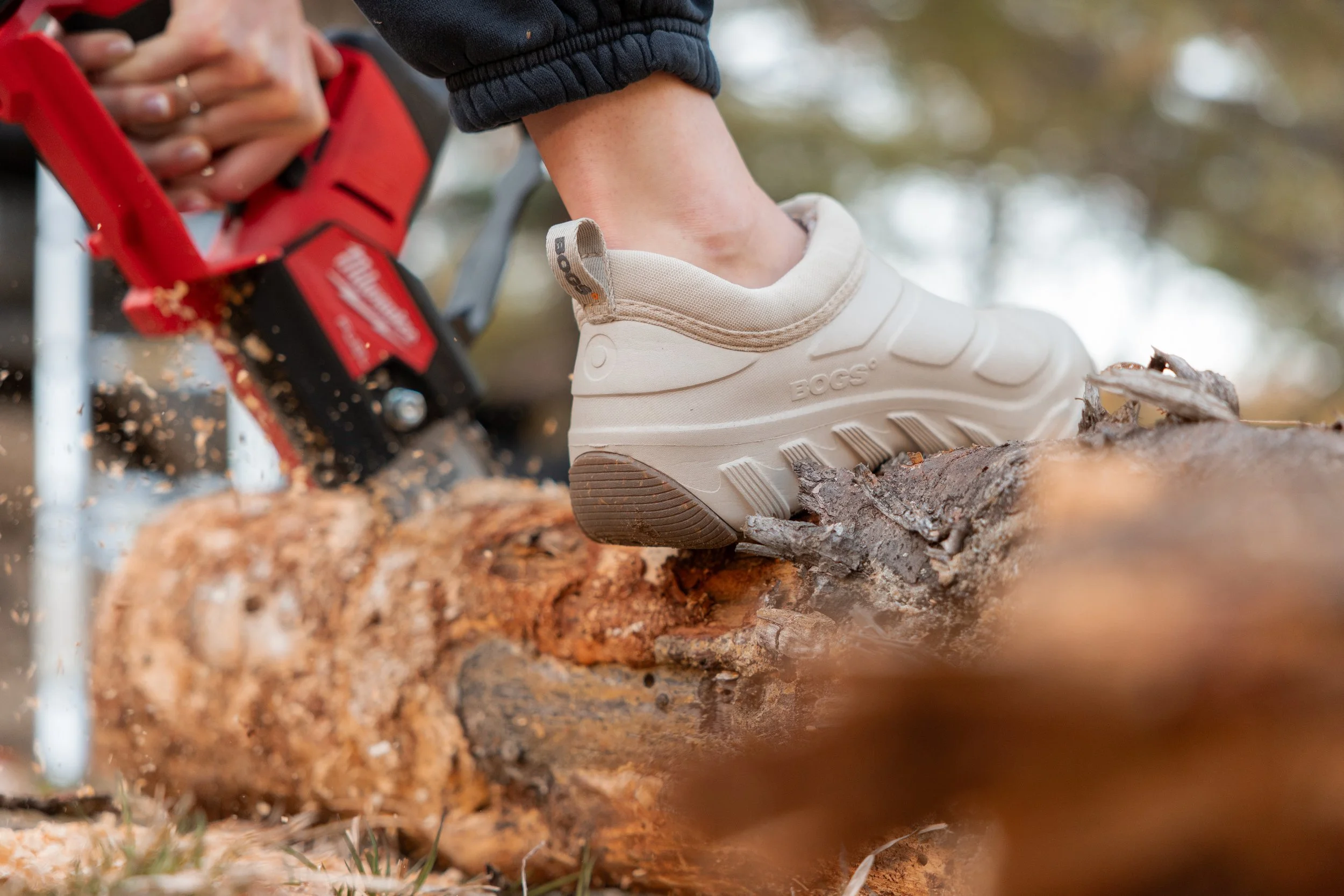 Close-up of a person's foot in a beige BOGS waterproof boot stepping on a log outdoors, with a red chainsaw cutting the log nearby.