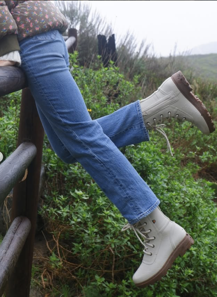 Person sitting on a wooden railing, wearing blue jeans and white boots, with their legs raised and the background showing green bushes and a foggy outdoor landscape.