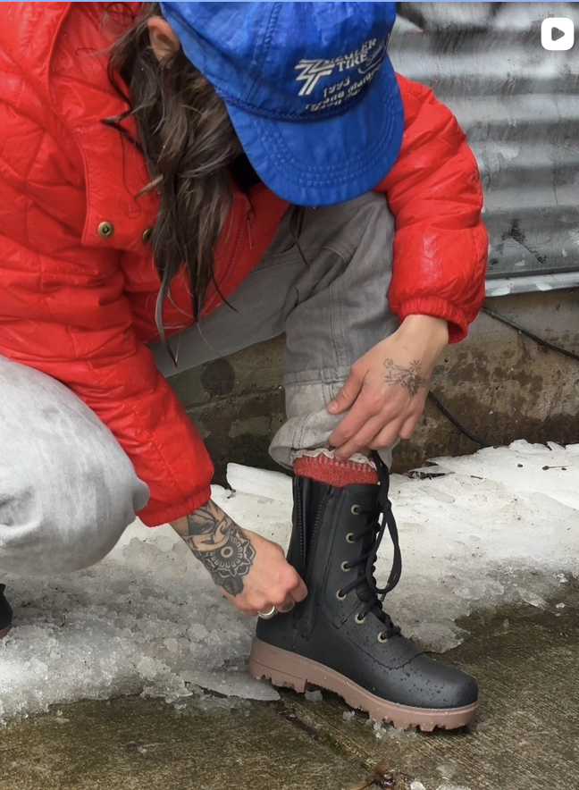 Person wearing a red jacket, blue cap, and gray pants is kneeling while putting on a black lace-up boot with a brown sole on icy ground.