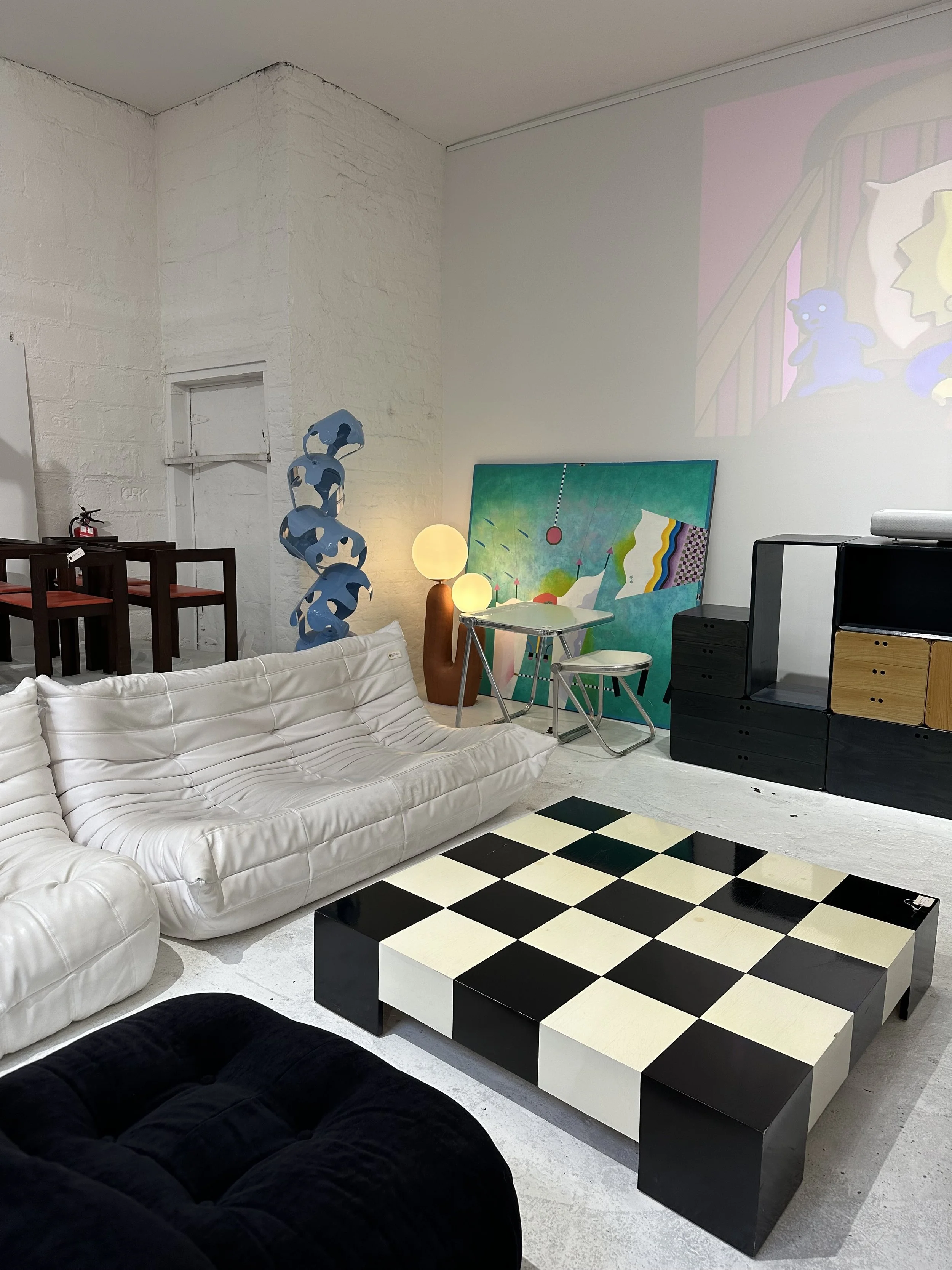 Interior of an art gallery or modern living space with various furniture pieces. Features include a white leather couch, black and white checkered table, black and wood storage unit, abstract blue sculpture, colorful artwork, and decorative lamps.