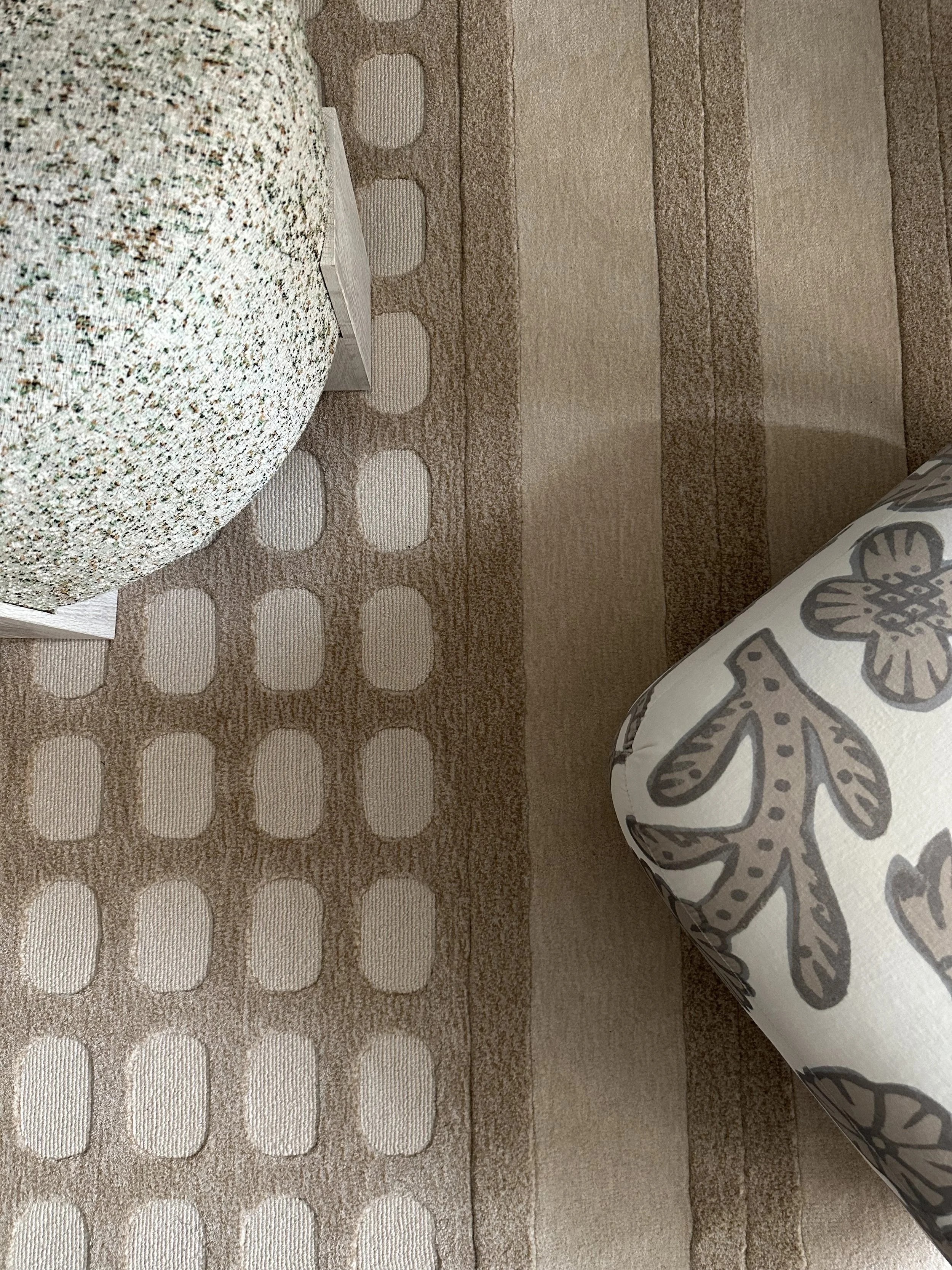 Close-up of a beige carpet with circular and striped patterns. Part of a bed with a floral patterned blanket is visible on the right, and a small wooden step stool is on the left.