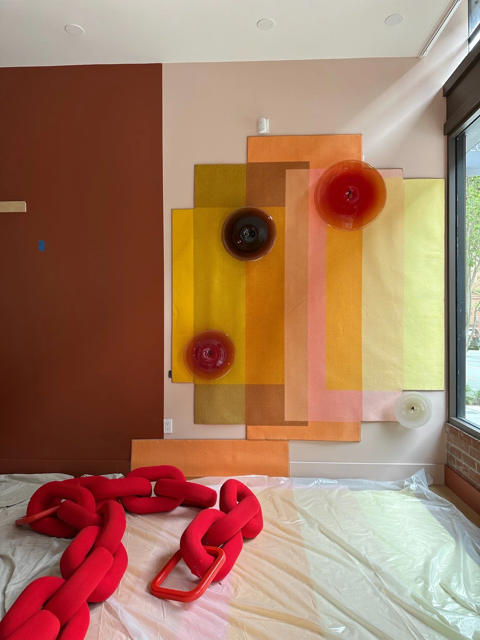 Interior wall with colorful modern art installation, featuring multicolored fabric panels and several large red and brown sculptural elements placed on a bed covered with plastic and red tubular objects.