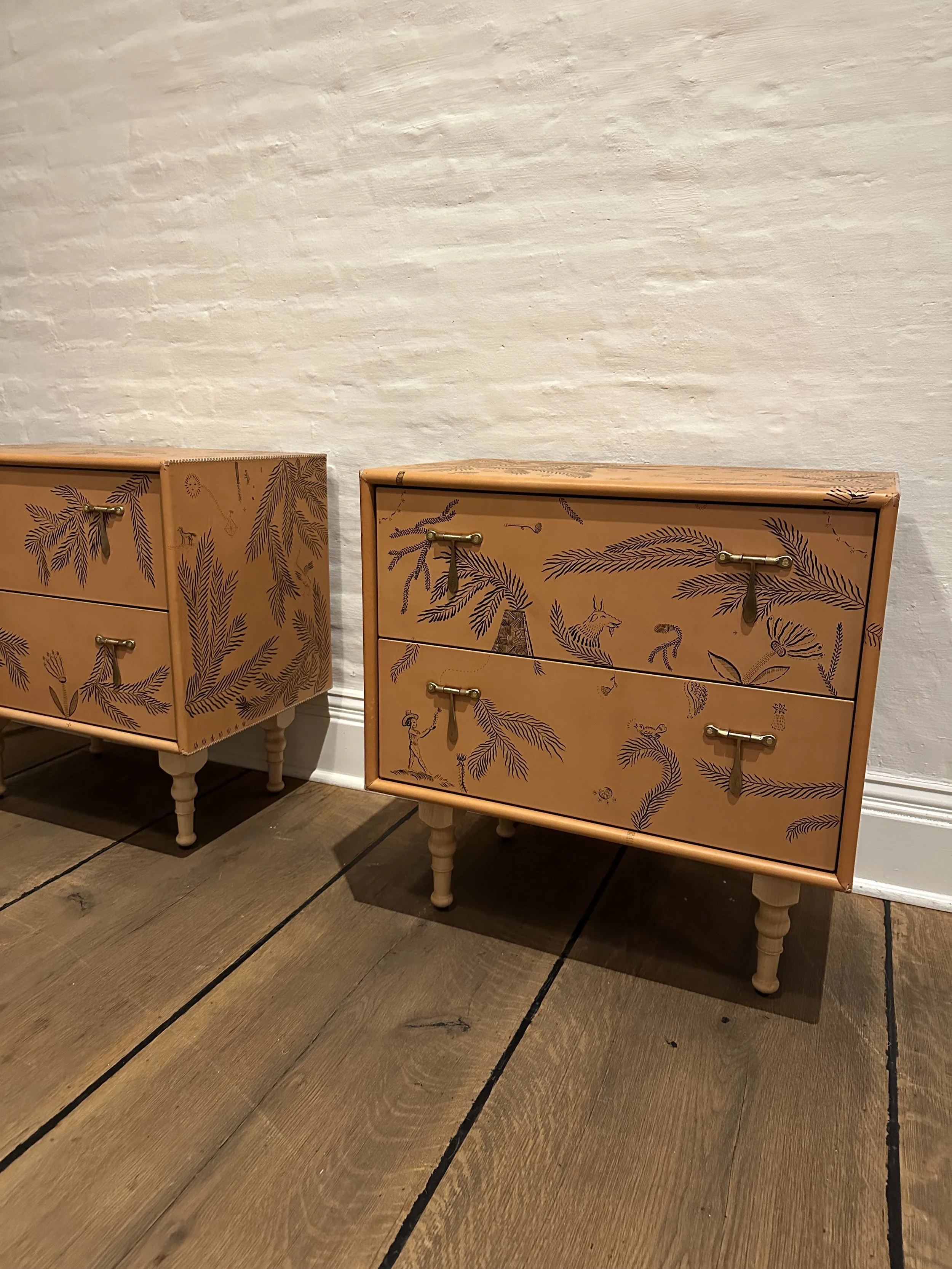Two peach-colored wooden dressers with botanical and animal illustrations, brass handles, and turned legs placed against a white brick wall on a wooden floor.
