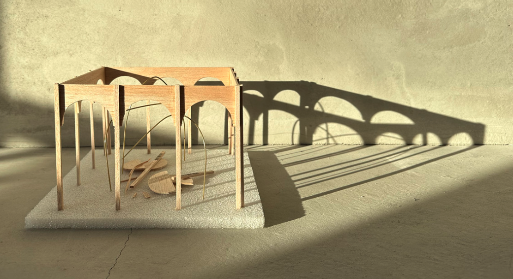 Wooden architectural model of an arched building casting shadows on a concrete surface.