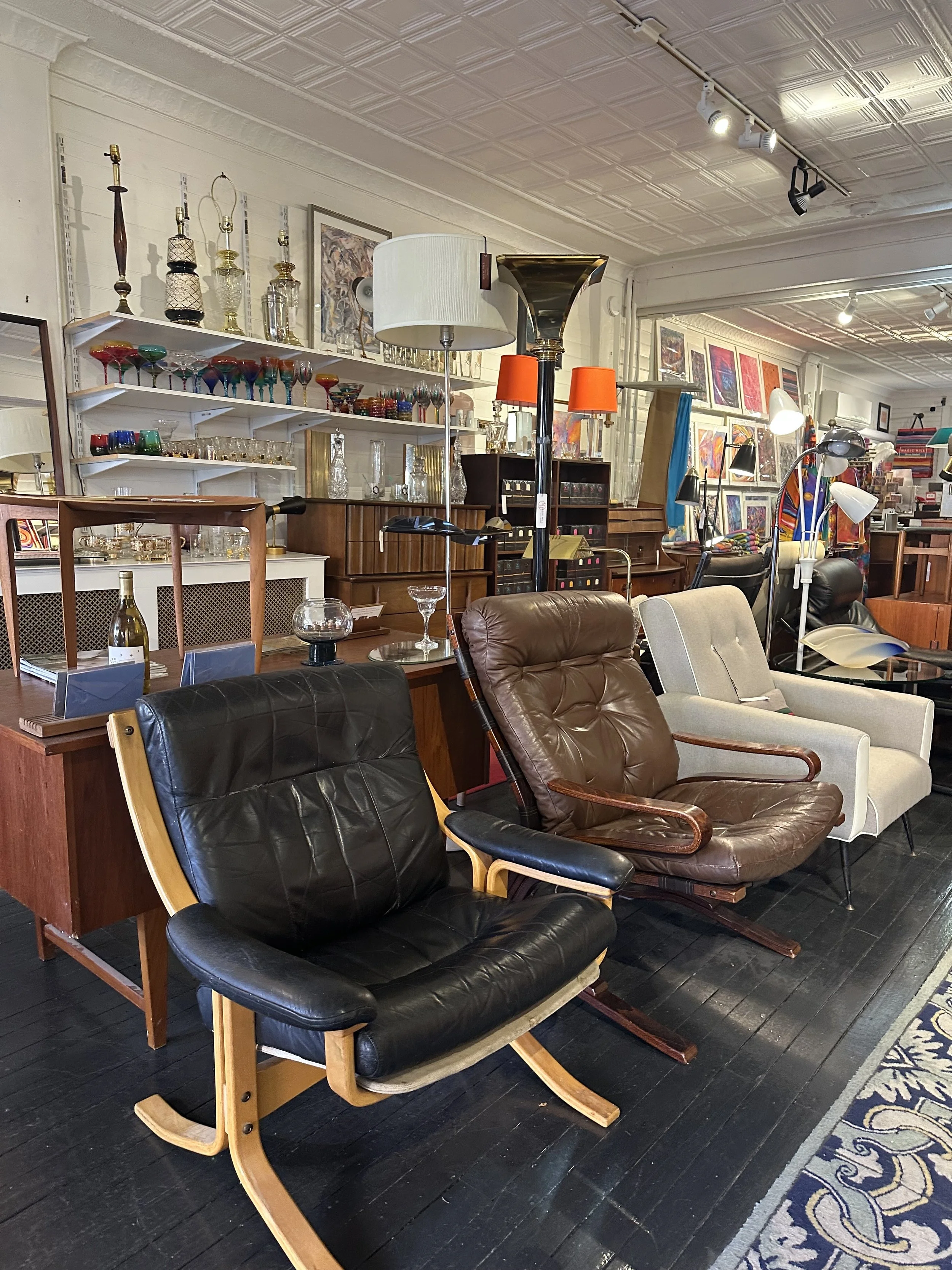 Various vintage furniture pieces including rocking chairs, lamps, and shelving units in a store with artwork and glassware displayed on shelves.