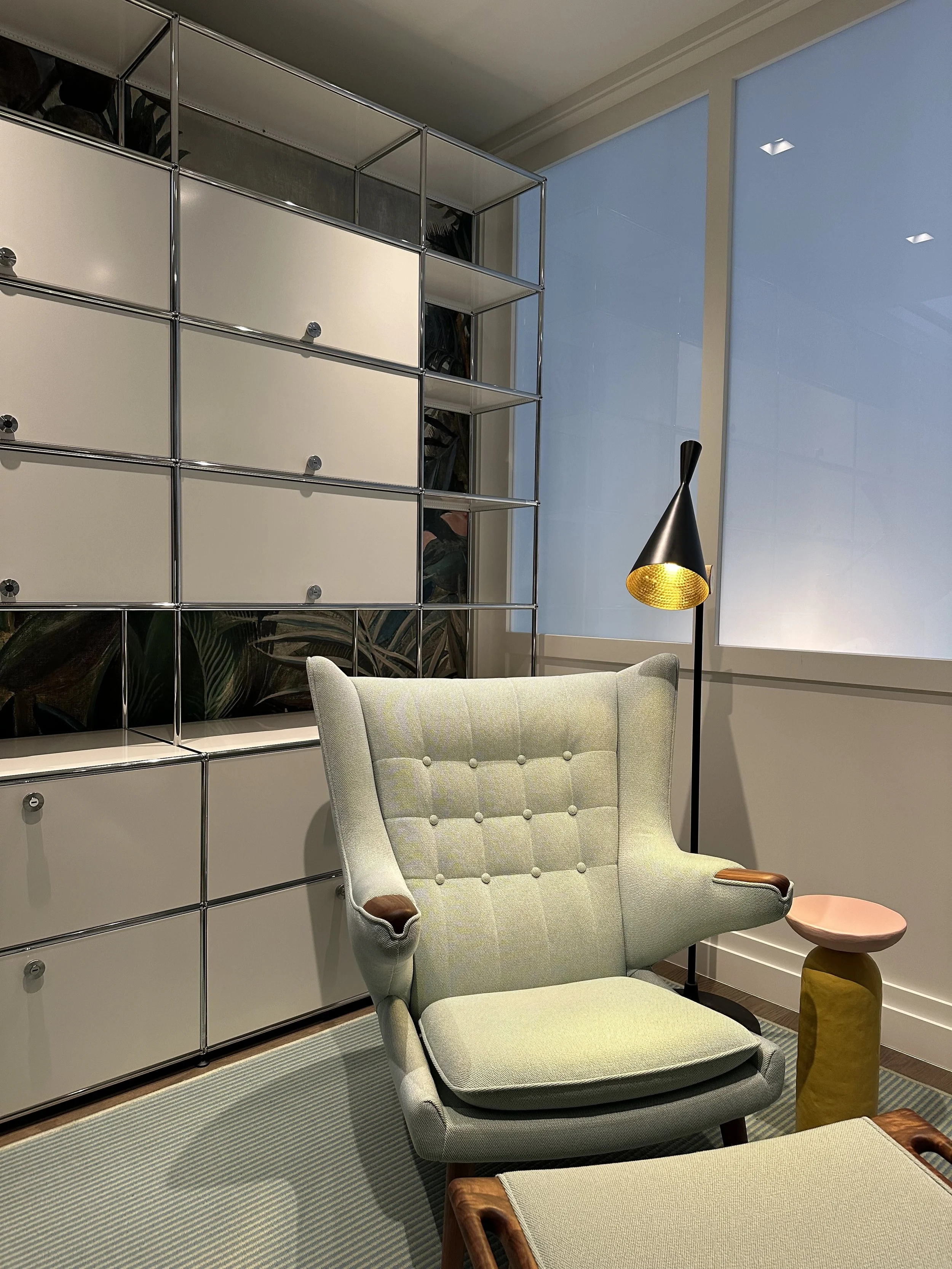A beige armchair with tufted backrest and wooden accents, a small pink side table, a black and yellow floor lamp, and a metallic and white storage cabinet with multiple drawers, in a room with large windows and a patterned carpet.