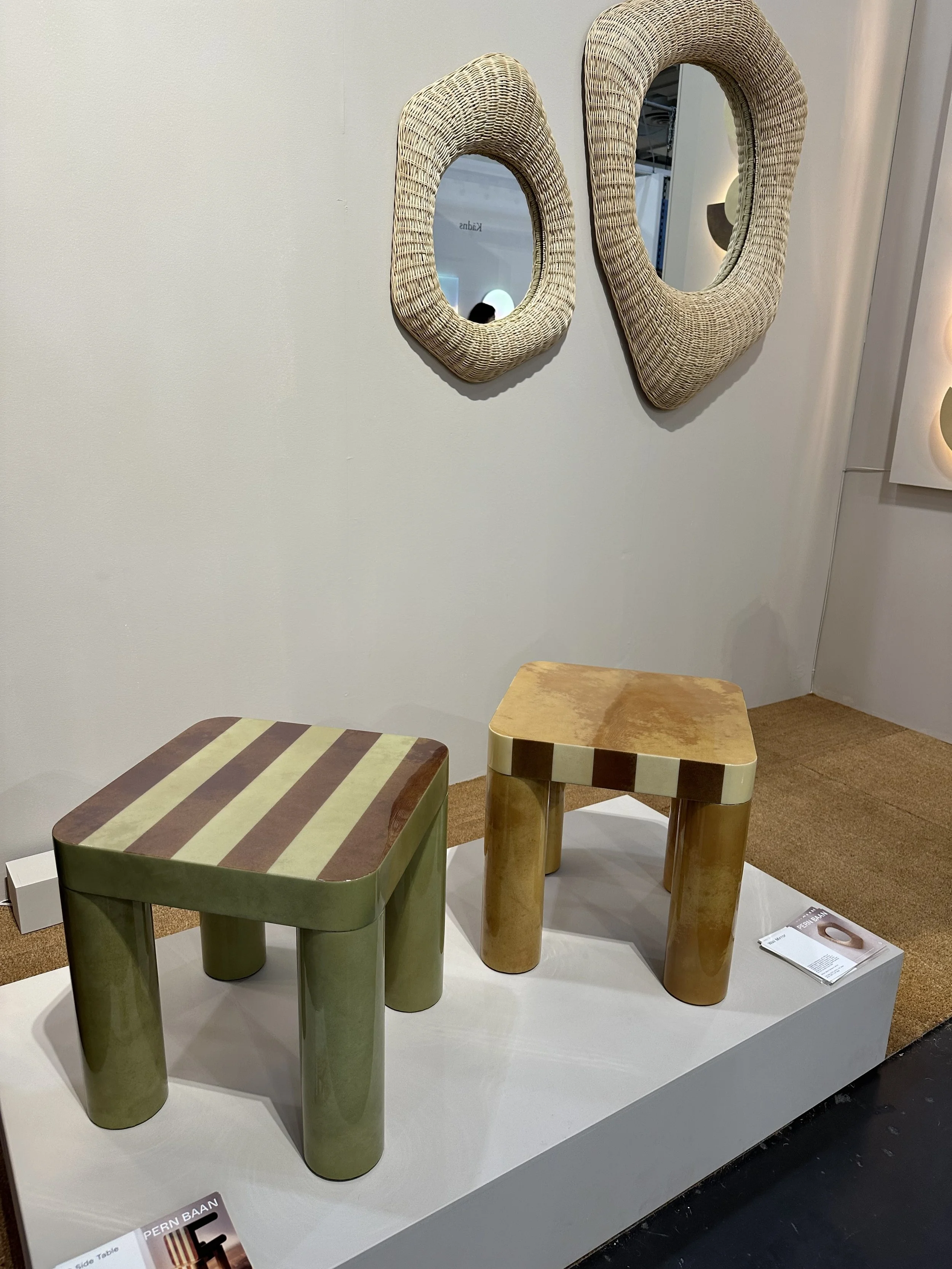 Two modern side tables made of wood, with one green and one beige, displayed on a white platform in a furniture store. In the background, two woven mirrors are mounted on a white wall.