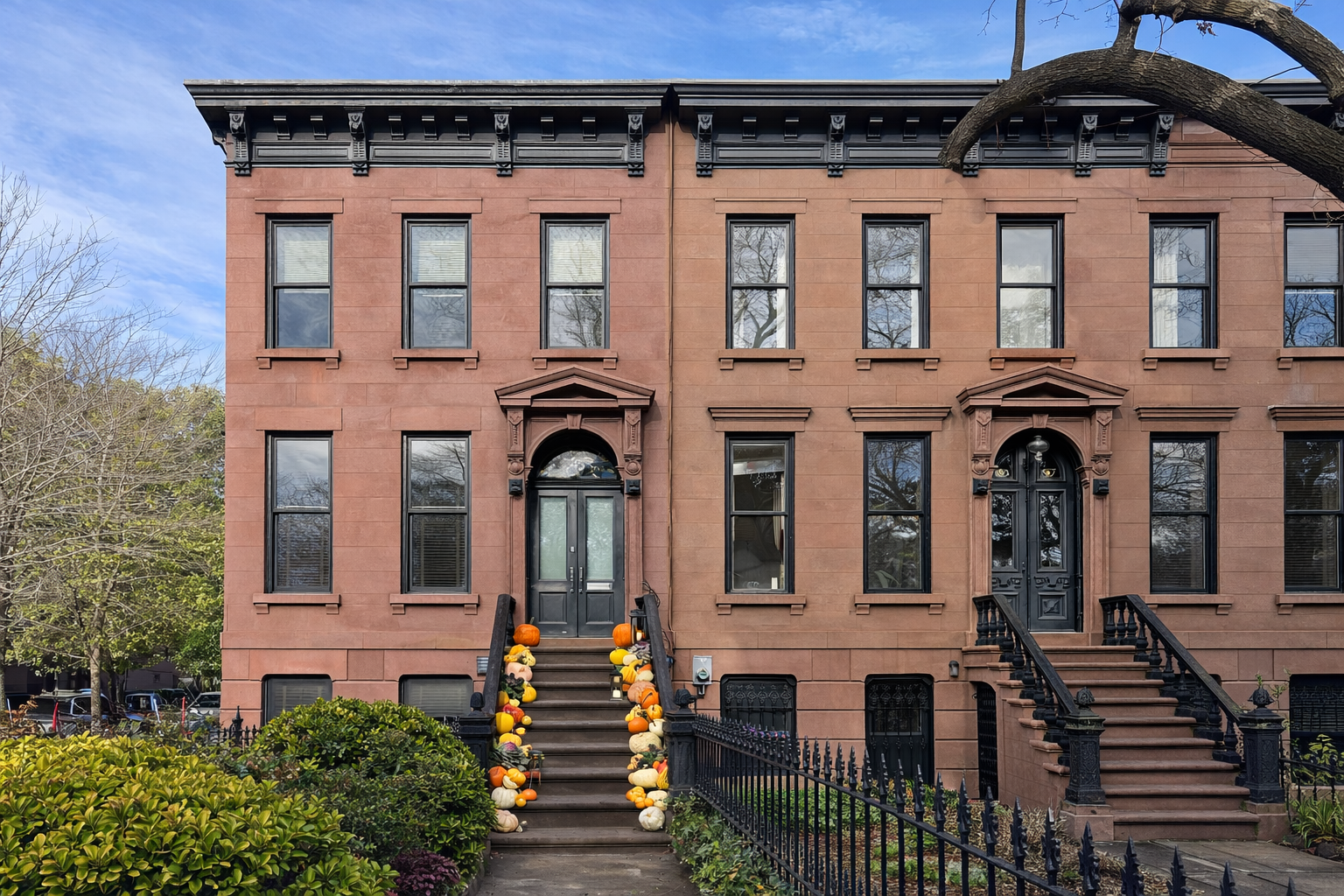 A brownstone townhouse with two entrances, decorated with pumpkins, fall decorations, and black iron fences, with trees and greenery in front.