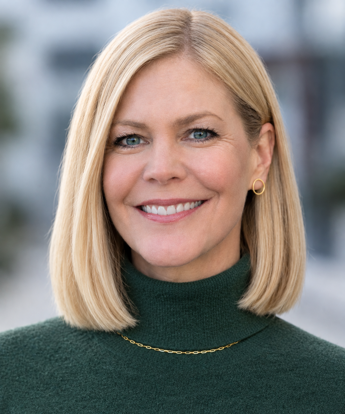 A smiling woman with blonde hair and blue eyes wearing a dark green turtleneck sweater, a gold chain necklace, and gold hoop earrings.