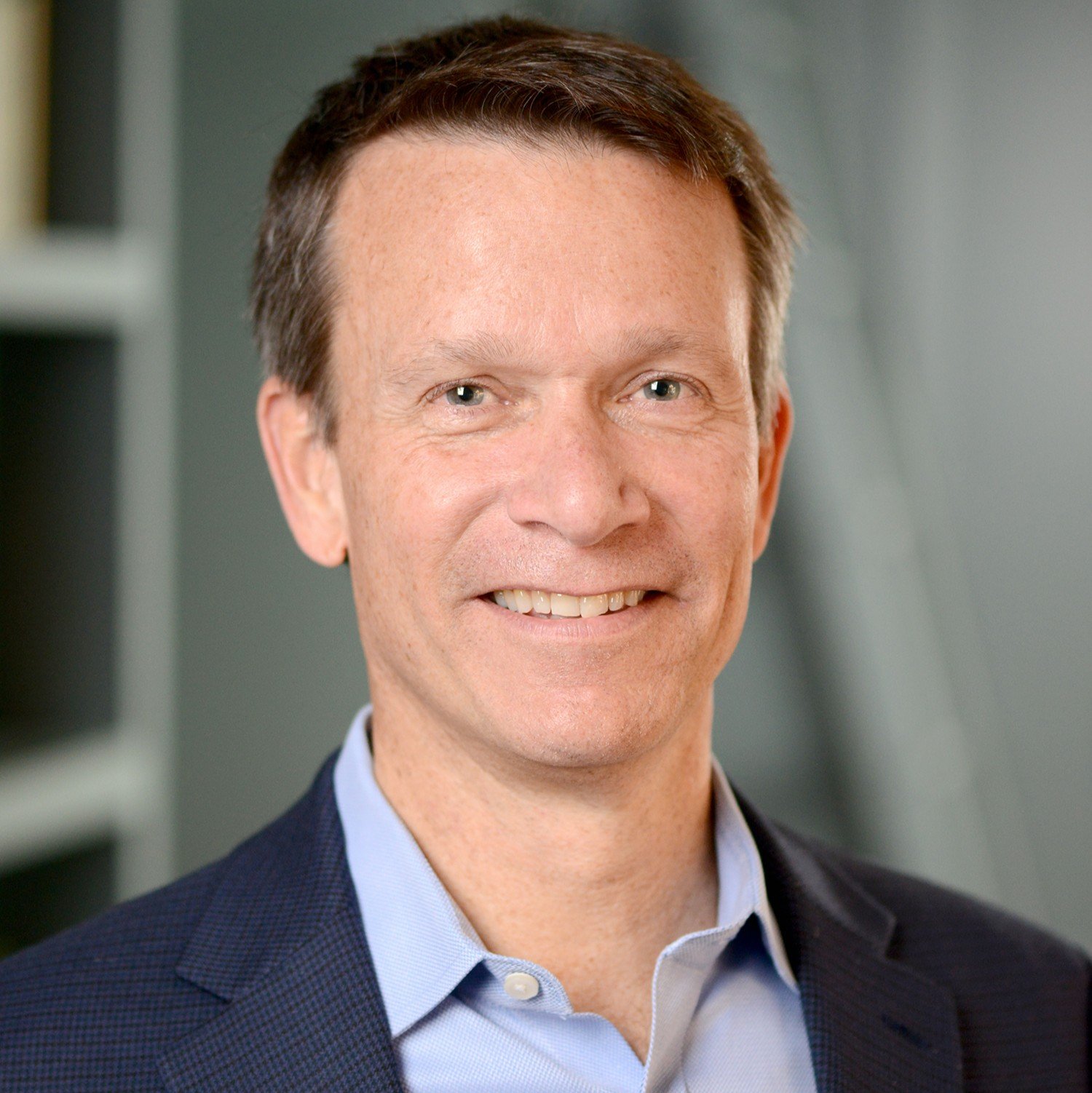 Portrait of smiling man with short brown hair, light blue eyes, wearing a dark blue blazer and light blue shirt.