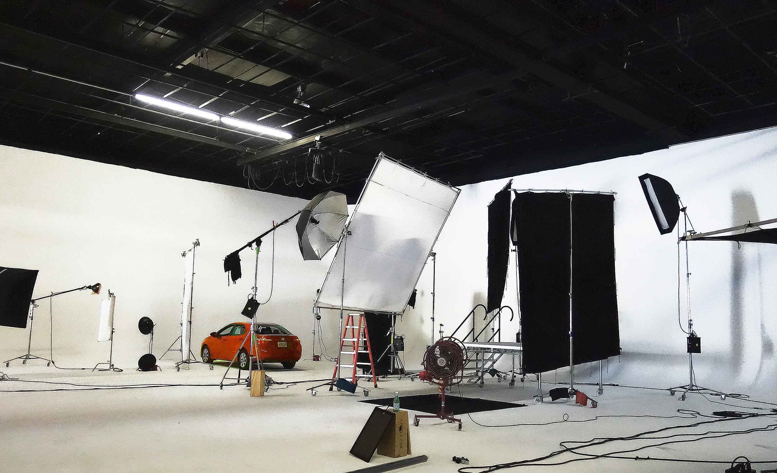Photo of a photography studio with lighting equipment, backdrop, and a small orange car prop, with various stands and tools around.