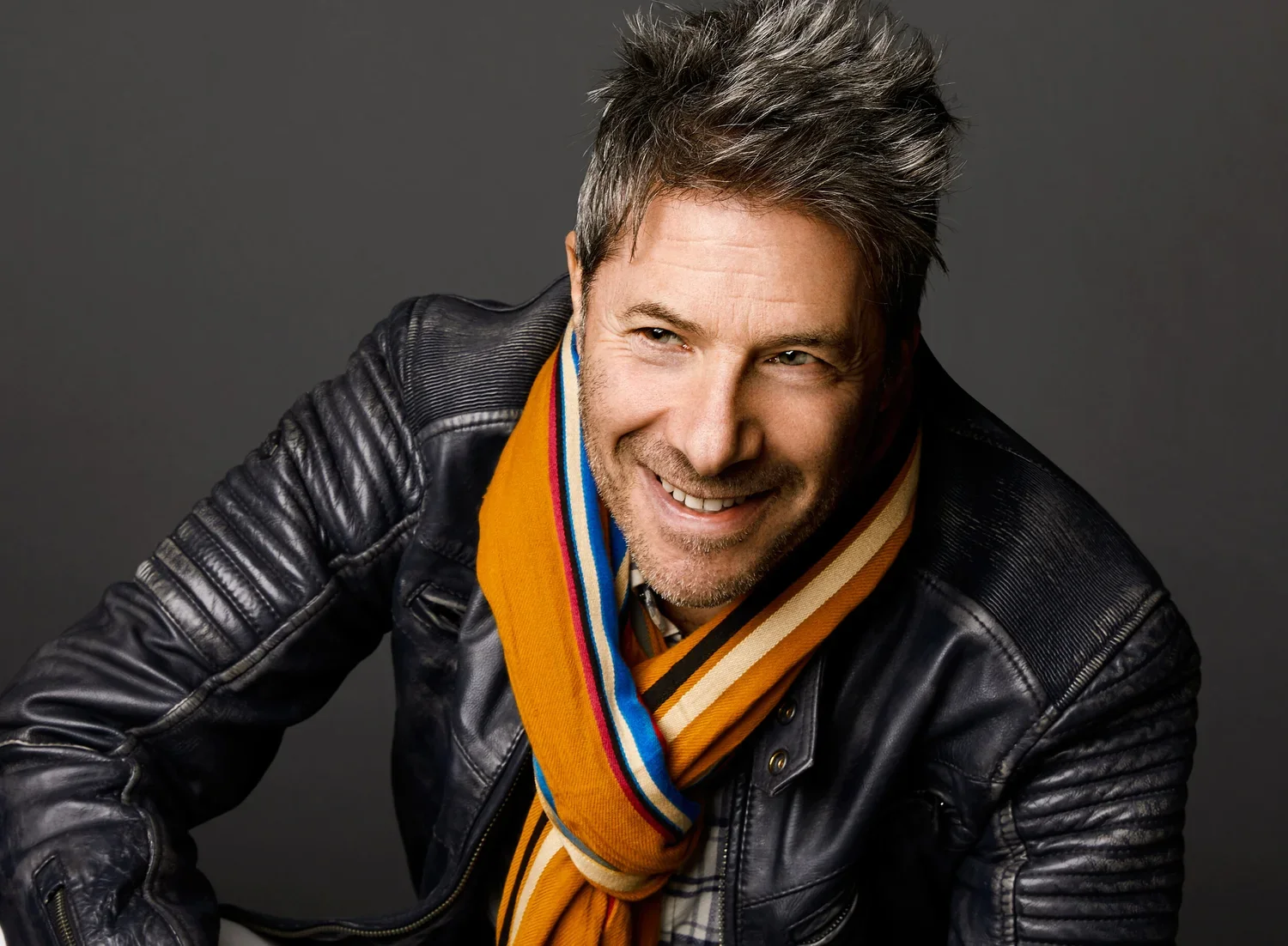 A man with short, spiky gray hair and a beard smiling, wearing a black leather jacket and a colorful striped scarf against a dark background.