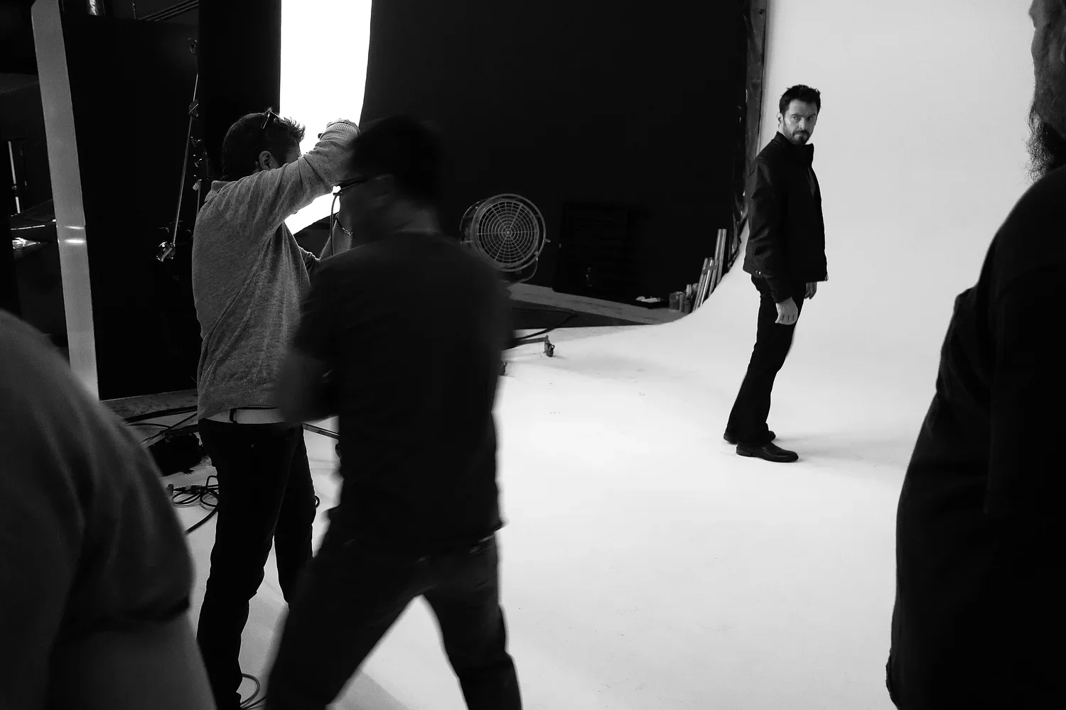 Black and white photo of a photoshoot in a studio. A man stands on a white backdrop, facing slightly to the right, while several crew members adjust equipment around him.