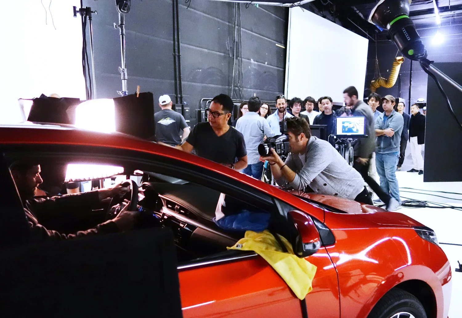 A behind-the-scenes scene of a car commercial or film shoot with a red car, a driver at the wheel, and a group of people including camera operators and crew members filming and observing inside a studio with black walls and white background.