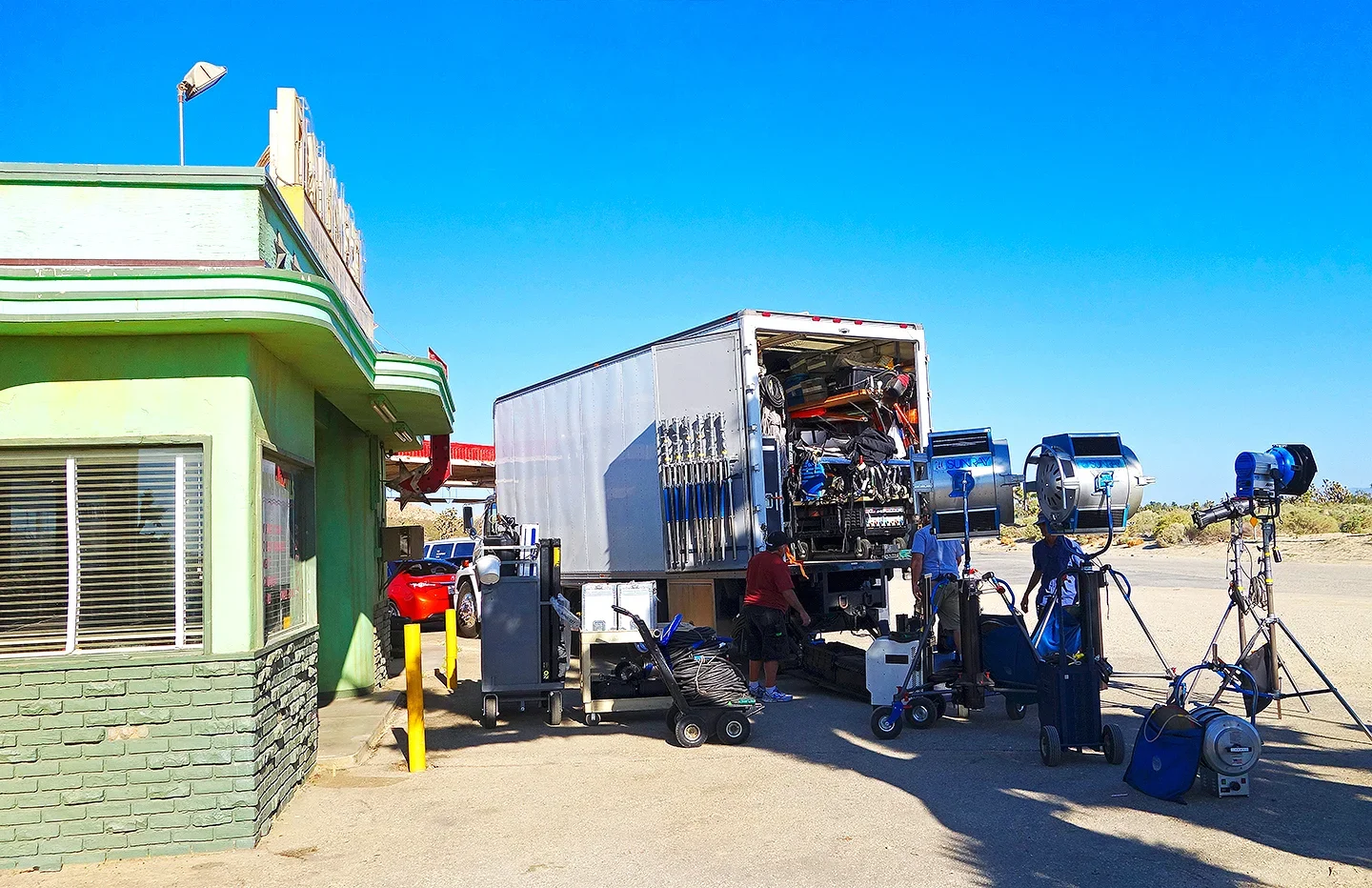 Film crew setting up equipment outside a green building on a sunny day, with camera lights and a truck holding gear.