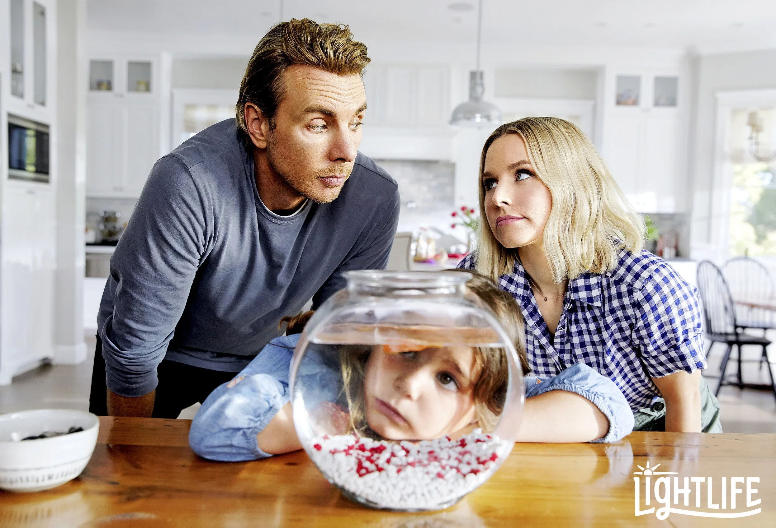 A young girl with red hair resting her head on a table in front of a fishbowl with a goldfish. Two adults observe her with concern in a bright kitchen. The LightLife logo is in the bottom right corner.