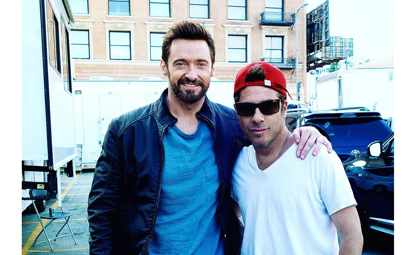Two men posing together outdoors in a parking lot, one with a beard and leather jacket, and the other wearing sunglasses, a red cap, and a white t-shirt.