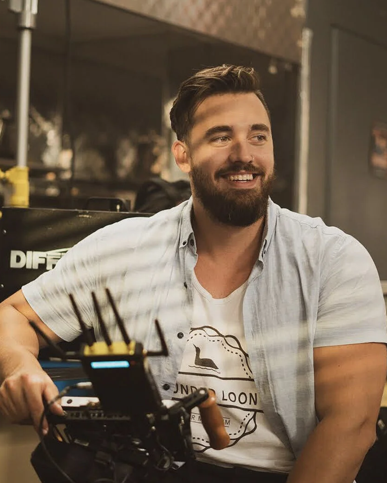 A smiling man with a beard and short dark hair, wearing a white t-shirt with a logo and a light blue open shirt, sitting indoors with a camera in the foreground.