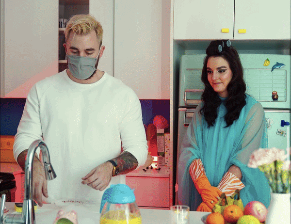 A man wearing a gray mask and white shirt standing at a kitchen counter with a woman in a blue dress with orange gloves, surrounded by fruits and kitchen items.