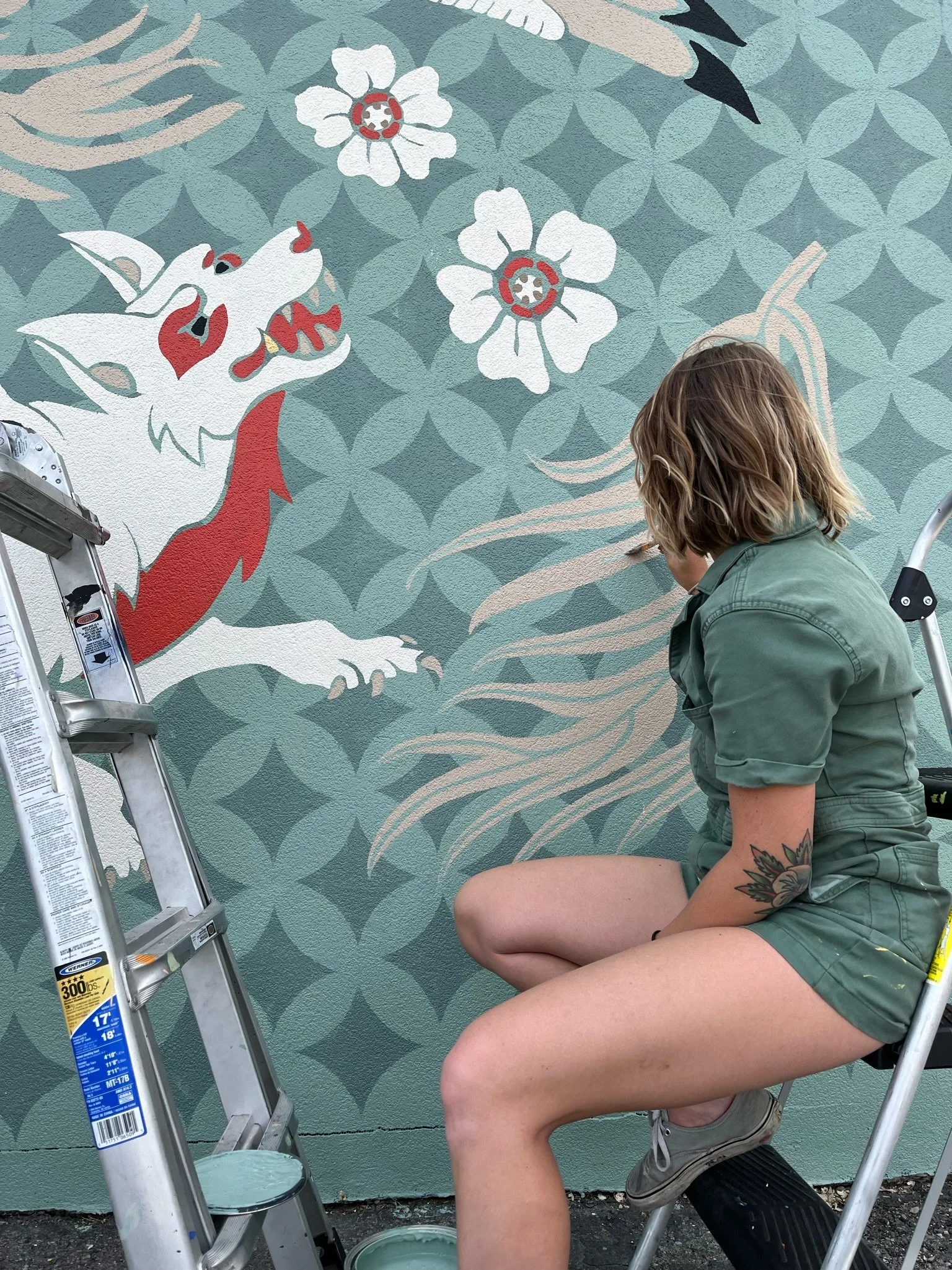 A person with shoulder-length hair and a tattoo on their arm is crouching in front of a mural of a dragon with white flowers and clouds. They are wearing a green jumpsuit and tan shoes, with a ladder and paint supplies nearby.