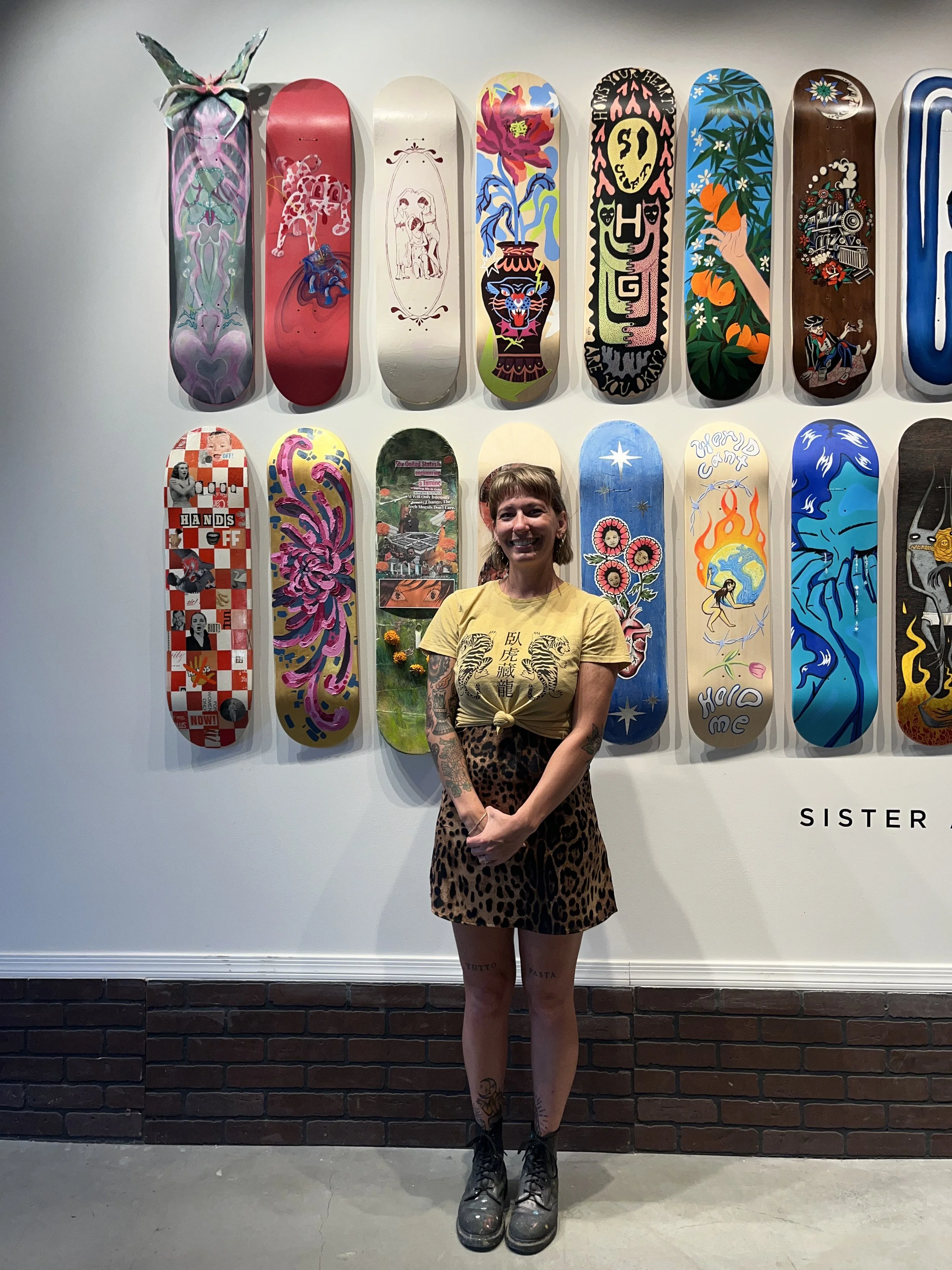 A woman with tattoos standing in front of a wall display of colorful skateboard decks with various designs including faces, animals, and abstract art.
