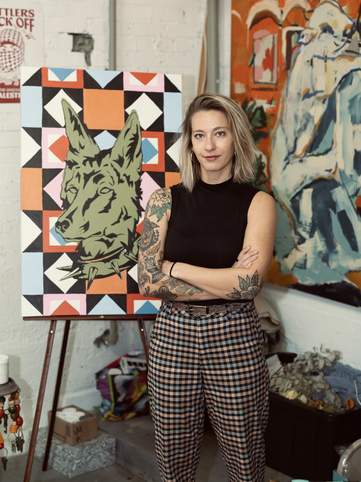 A woman with tattoos on her arms and blonde hair standing in front of colorful artwork in an art studio or gallery.