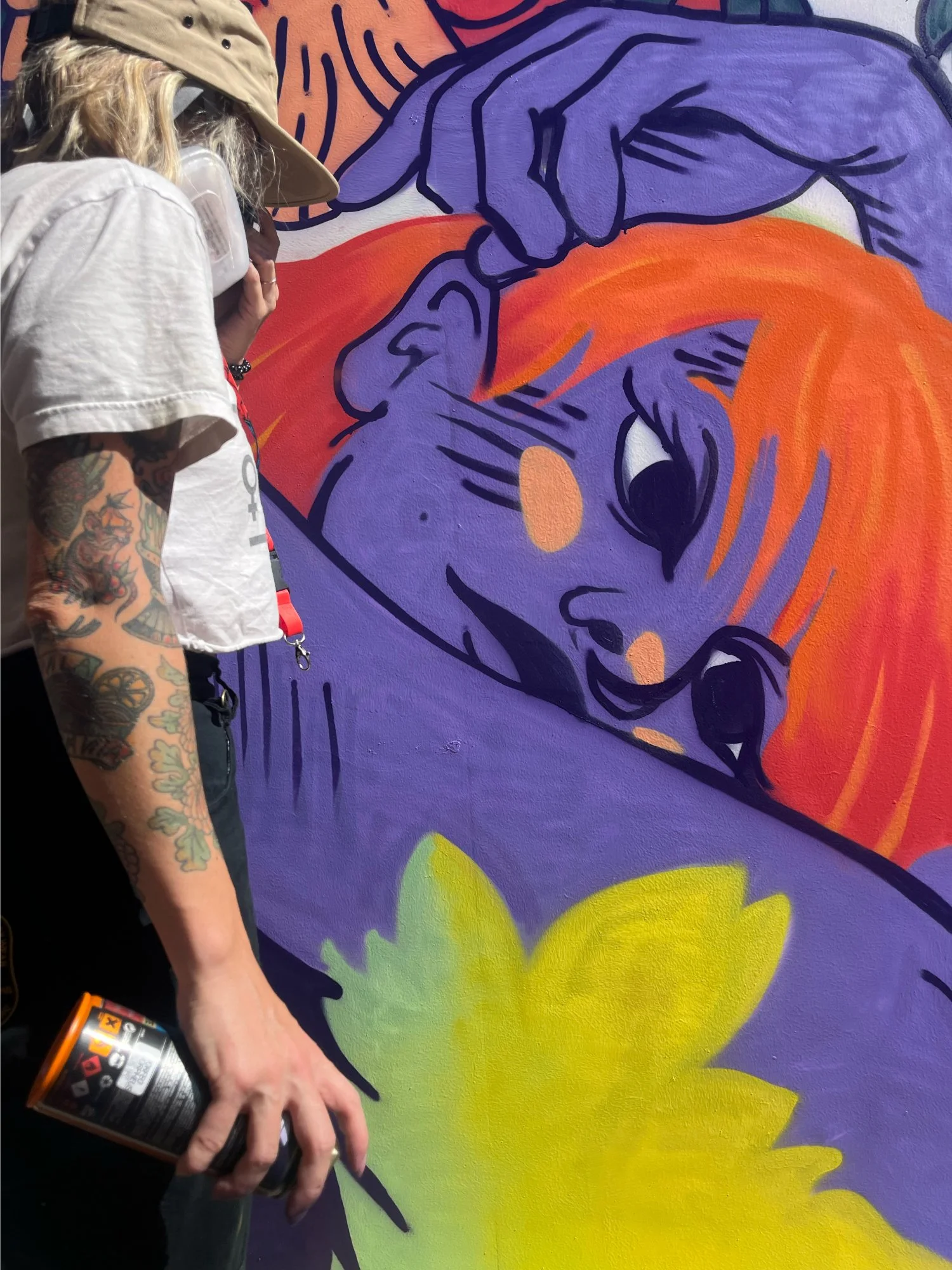 Person with long blonde hair and tattoos on arm, wearing a beige hat and white shirt, holding a spray paint can, working on a colorful mural that features purple, orange, yellow, and green sections with bold black outlines.