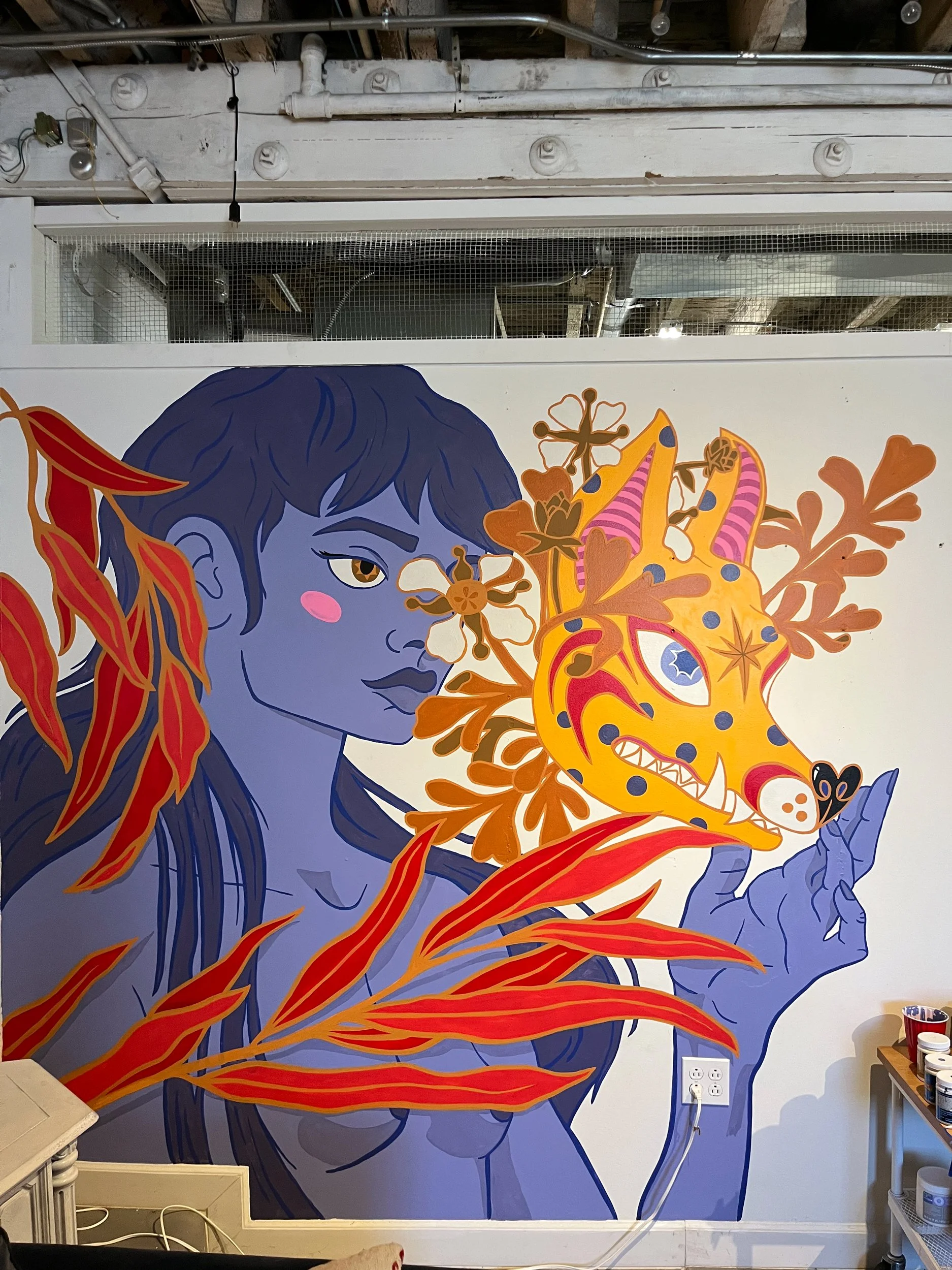 A colorful mural on a wall depicting a woman with dark blue hair and purple skin, surrounded by orange and pink foliage, with a stylized yellow and orange creature with blue spots.