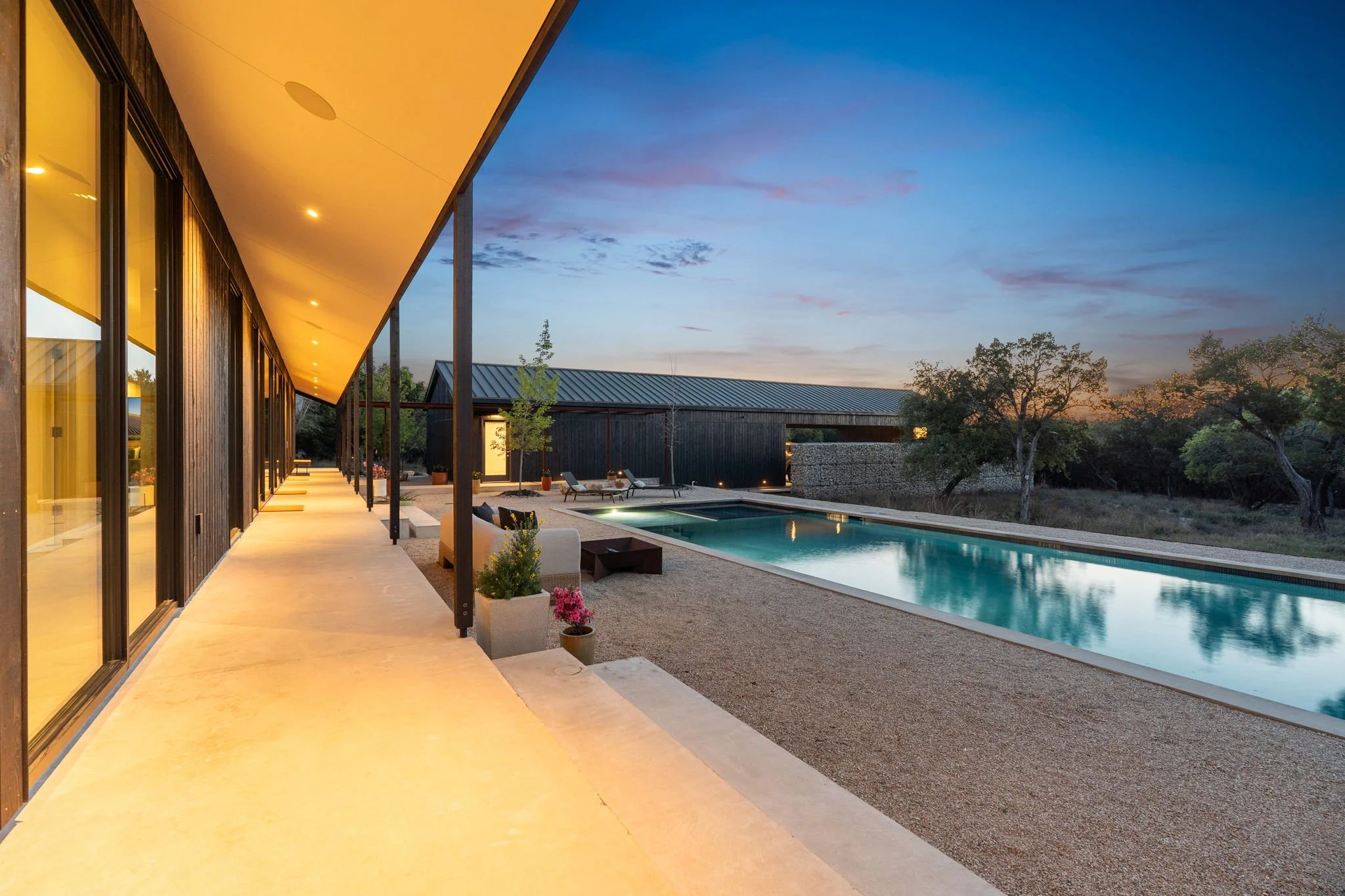 A modern house with a yellow-lit ceiling and large glass windows overlooking a backyard with a swimming pool, patio chairs, potted plants, and trees at sunset.