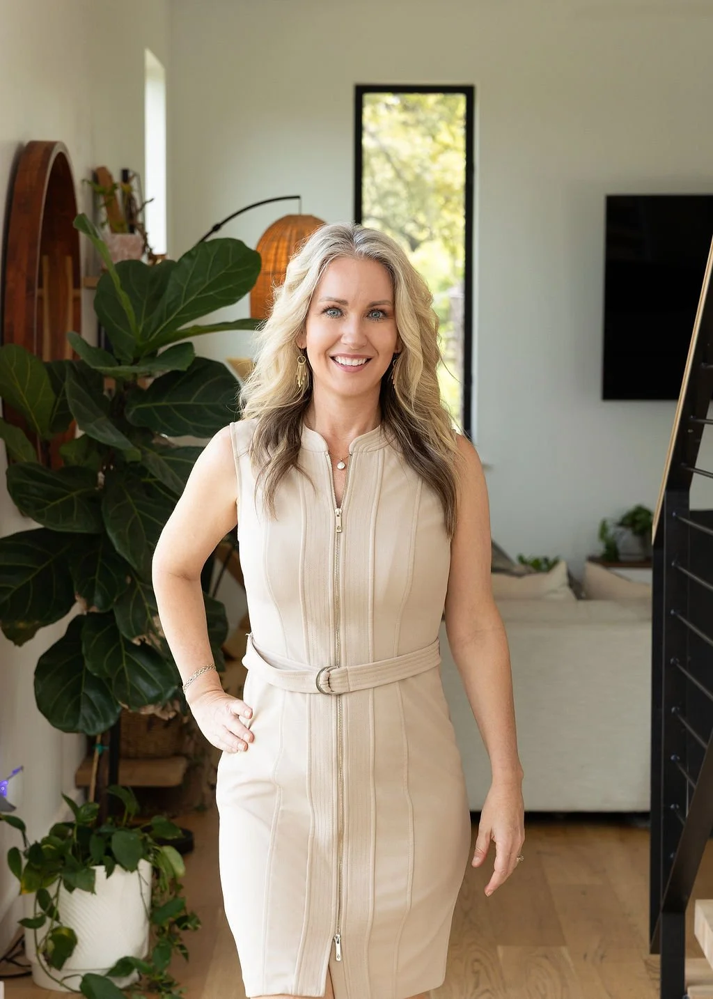 Austin Realtor Kristen Jacobs smiling in a beige sleeveless dress, standing indoors with green plants and natural light in the background.