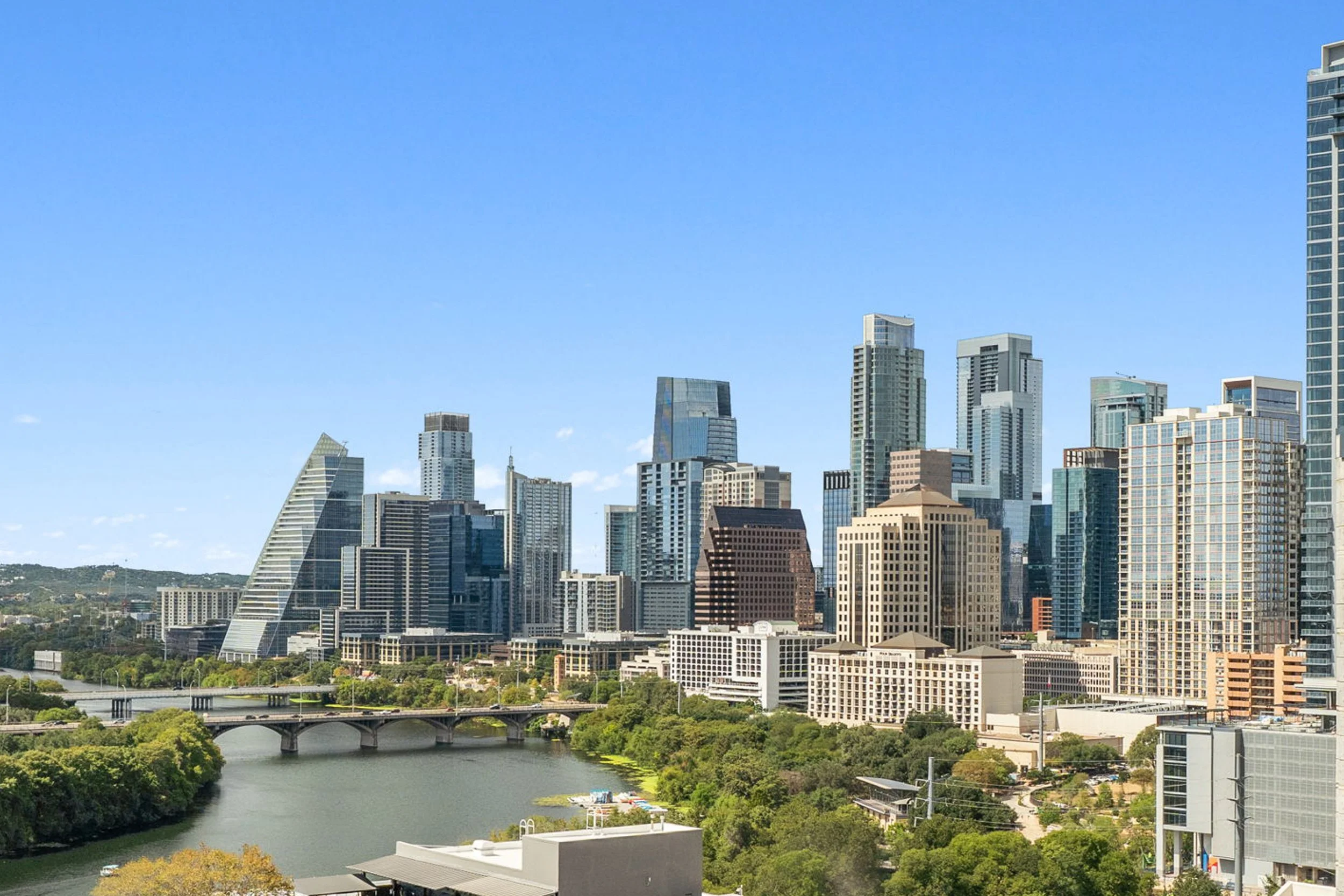 Downtown Austin, Texas skyline featuring modern skyscrapers, the Colorado River, and a bridge surrounded by green trees on a clear day.