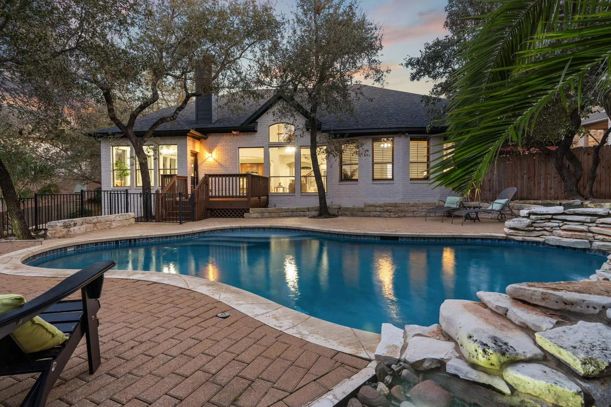 Backyard view of a house with lit windows, a wooden deck, trees, a swimming pool with lounge chairs, and a stacked stone feature in the evening.