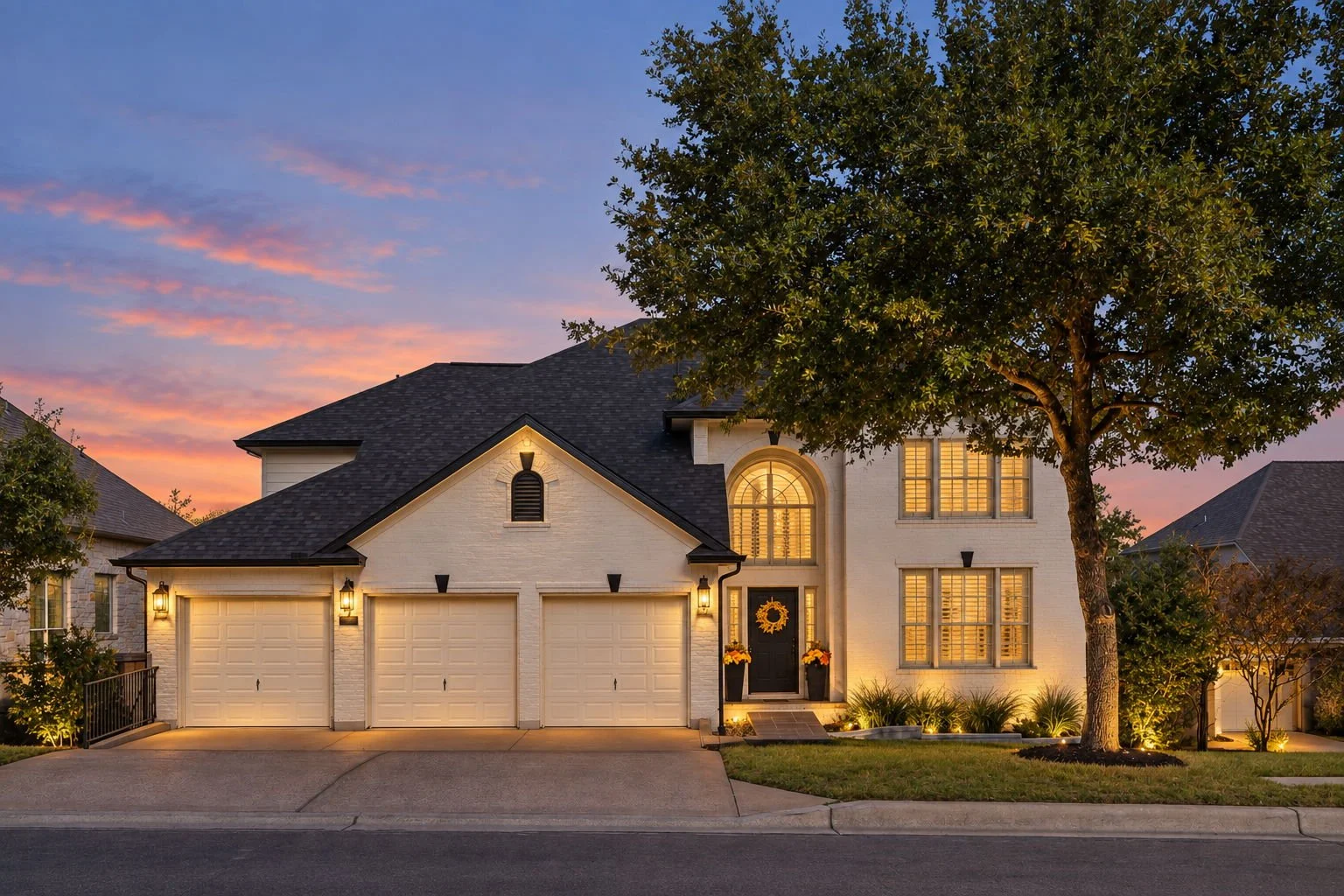 A modern two-story house with white brick exterior, three garage doors, and front porch with decorations. Illuminated by exterior lights at sunset with a large tree in the front yard.