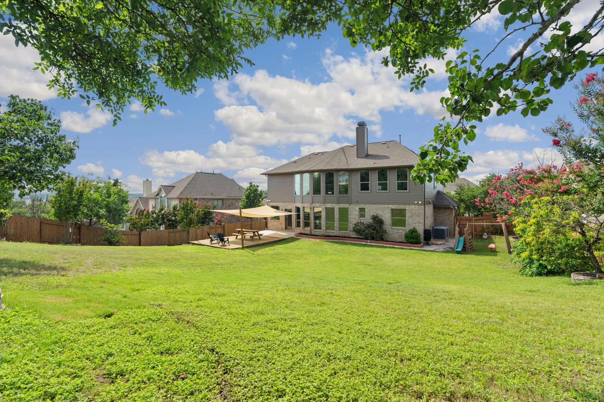 A backyard with a green lawn, trees, and a house with a deck. The sky is partly cloudy, and there are outdoor chairs, a small slide, and a play area.