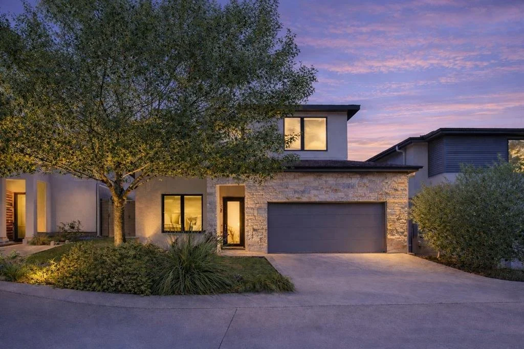 Modern two-story house with stone and stucco exterior, large trees, and a garage, illuminated during sunset.