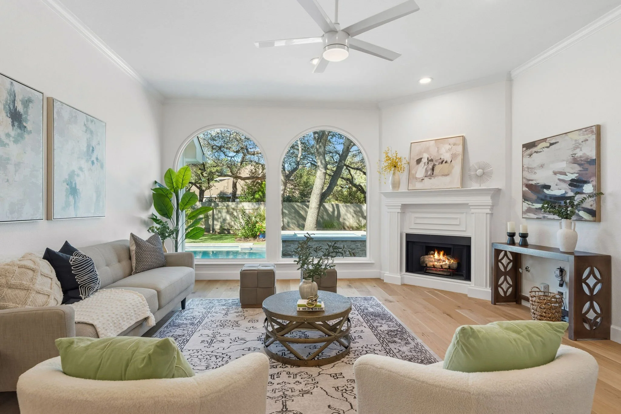 Bright living room with large arched windows, a white fireplace, a ceiling fan, a sofa with cushions, a coffee table, and wall art, overlooking a backyard with trees and a pool.