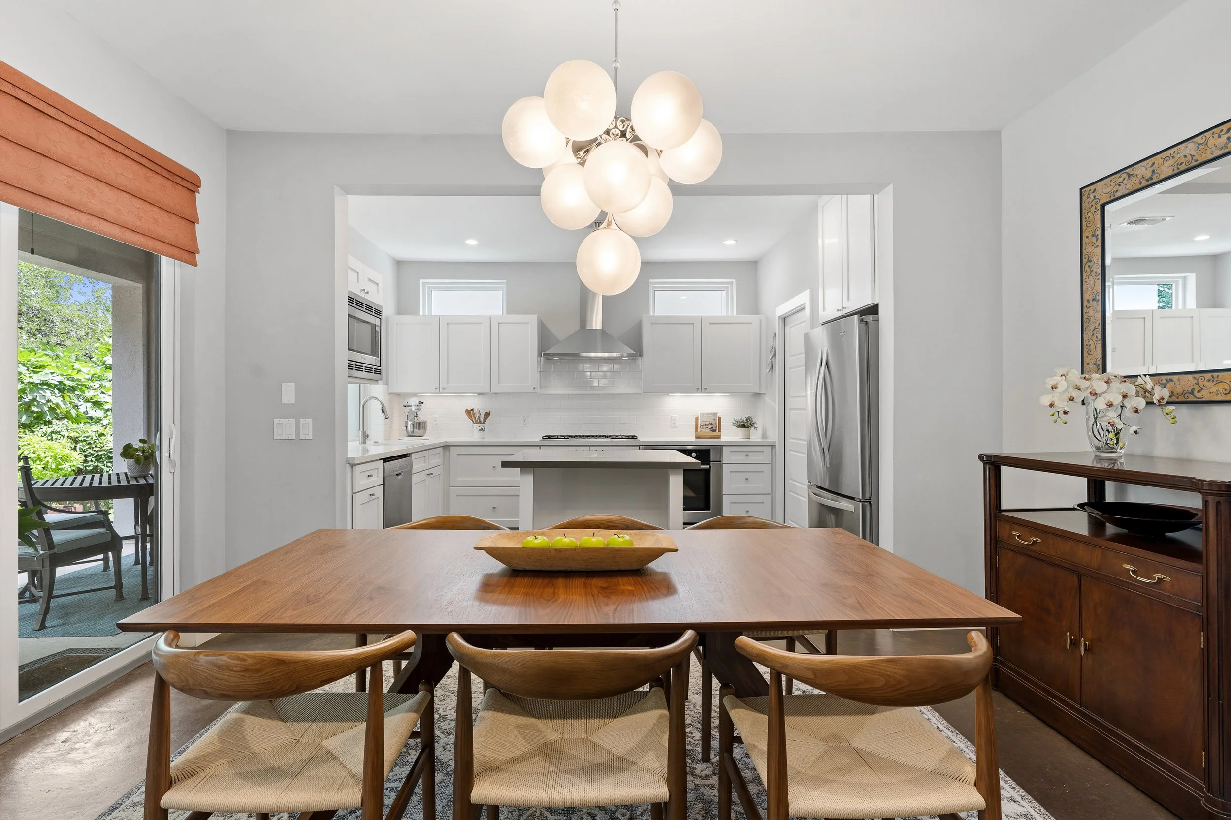 A modern kitchen and dining area with a wooden dining table and six matching chairs, a white kitchen with stainless steel appliances, and a large light fixture hanging from the ceiling. There is a sliding door leading outside on the left, and a mirror and a wooden side table with a vase of white flowers on the right.