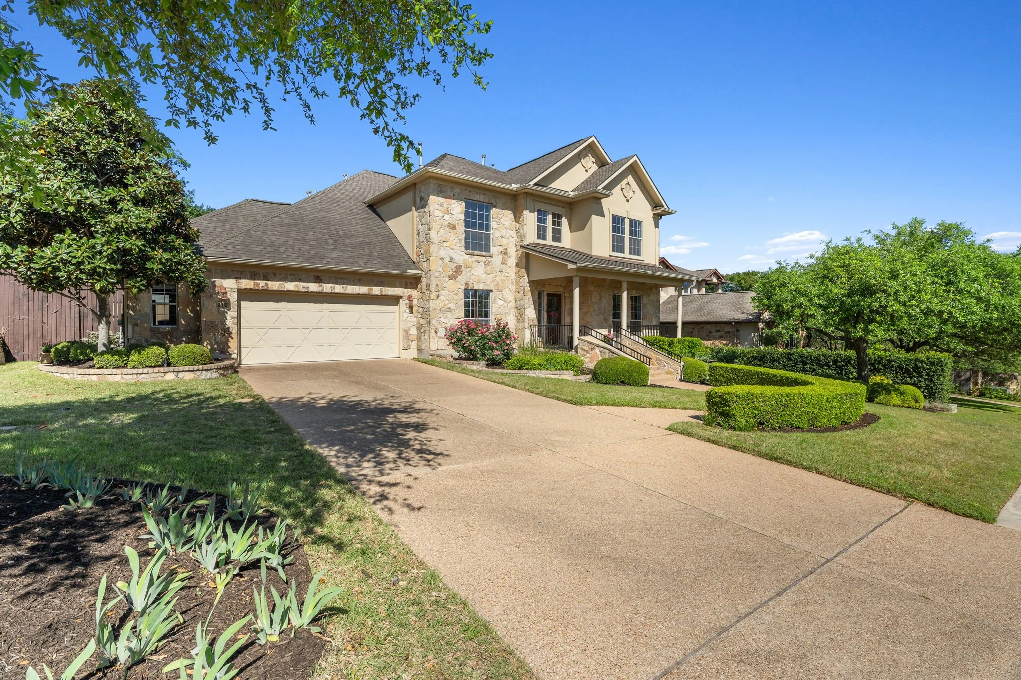 A two-story house with a stone facade, beige siding, and a gray shingle roof. It has a front porch with a black railing, several windows, and a closed garage door. The house is surrounded by a yard with neatly manicured bushes, trees, and flower beds, under a clear blue sky.