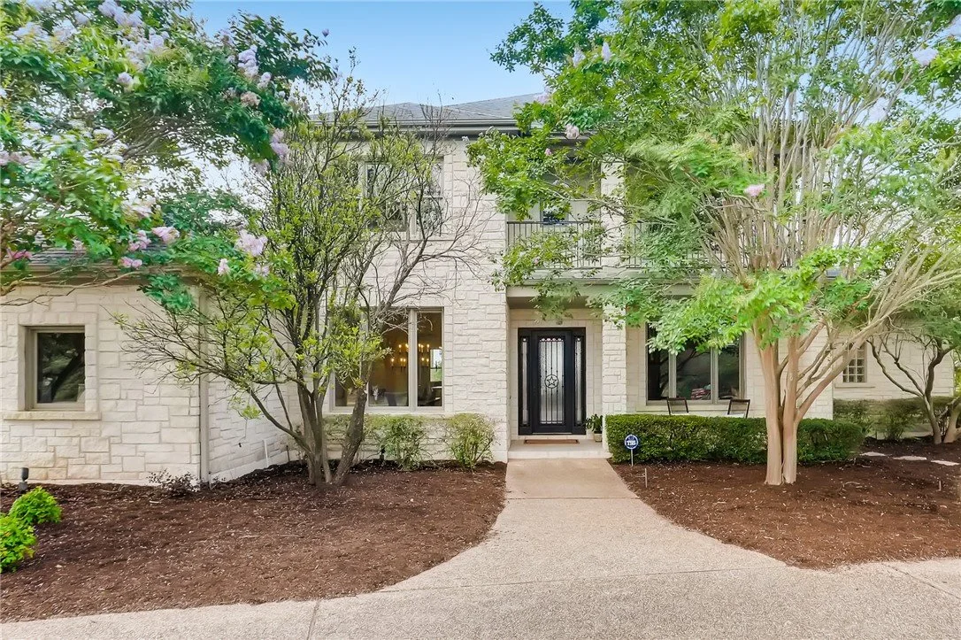 Front view of a two-story house with a black front door, stone exterior, and large windows, surrounded by trees and landscaped mulched garden beds.