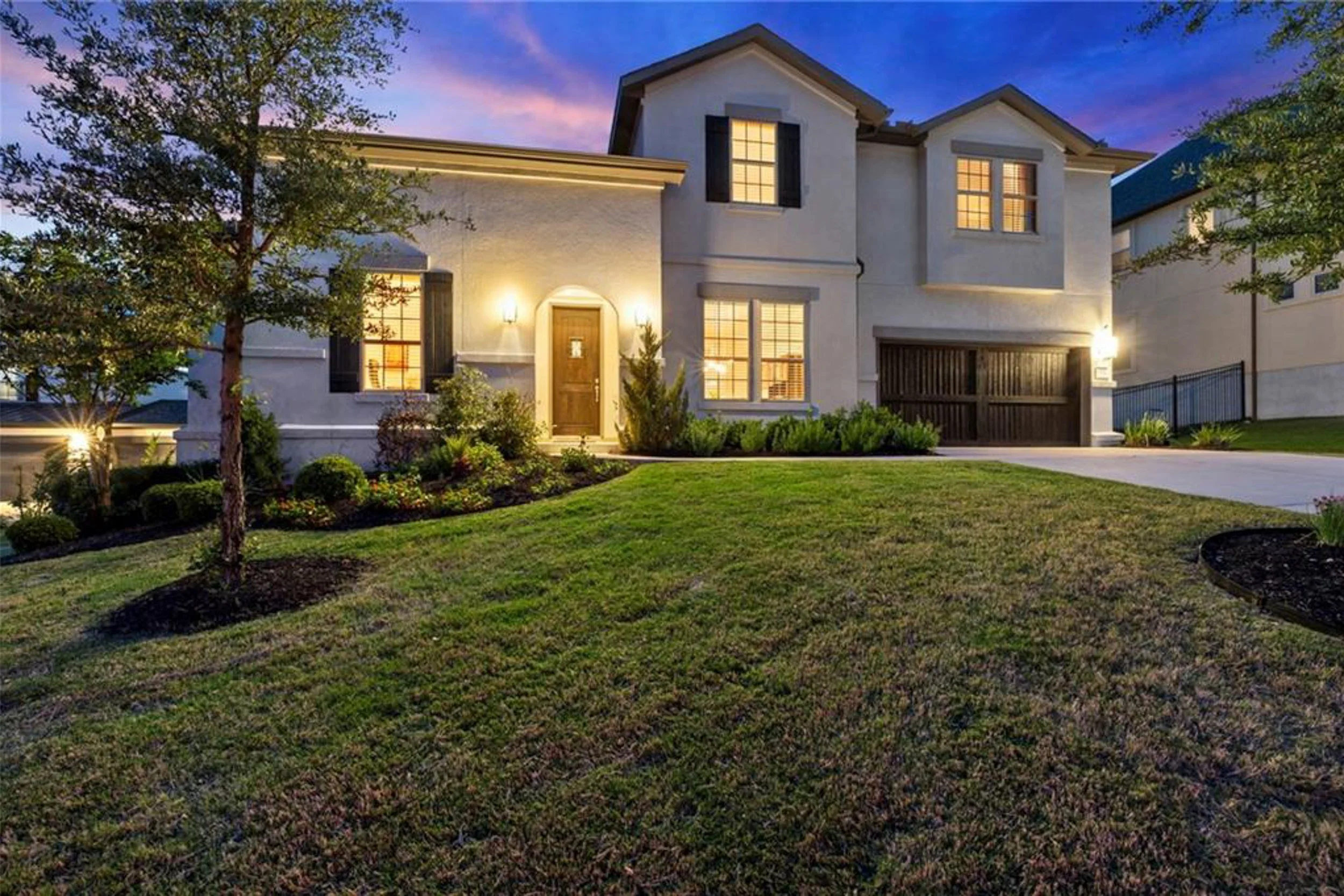 A two-story modern house with outdoor lighting, a manicured lawn, and trees, taken during early evening or dusk.