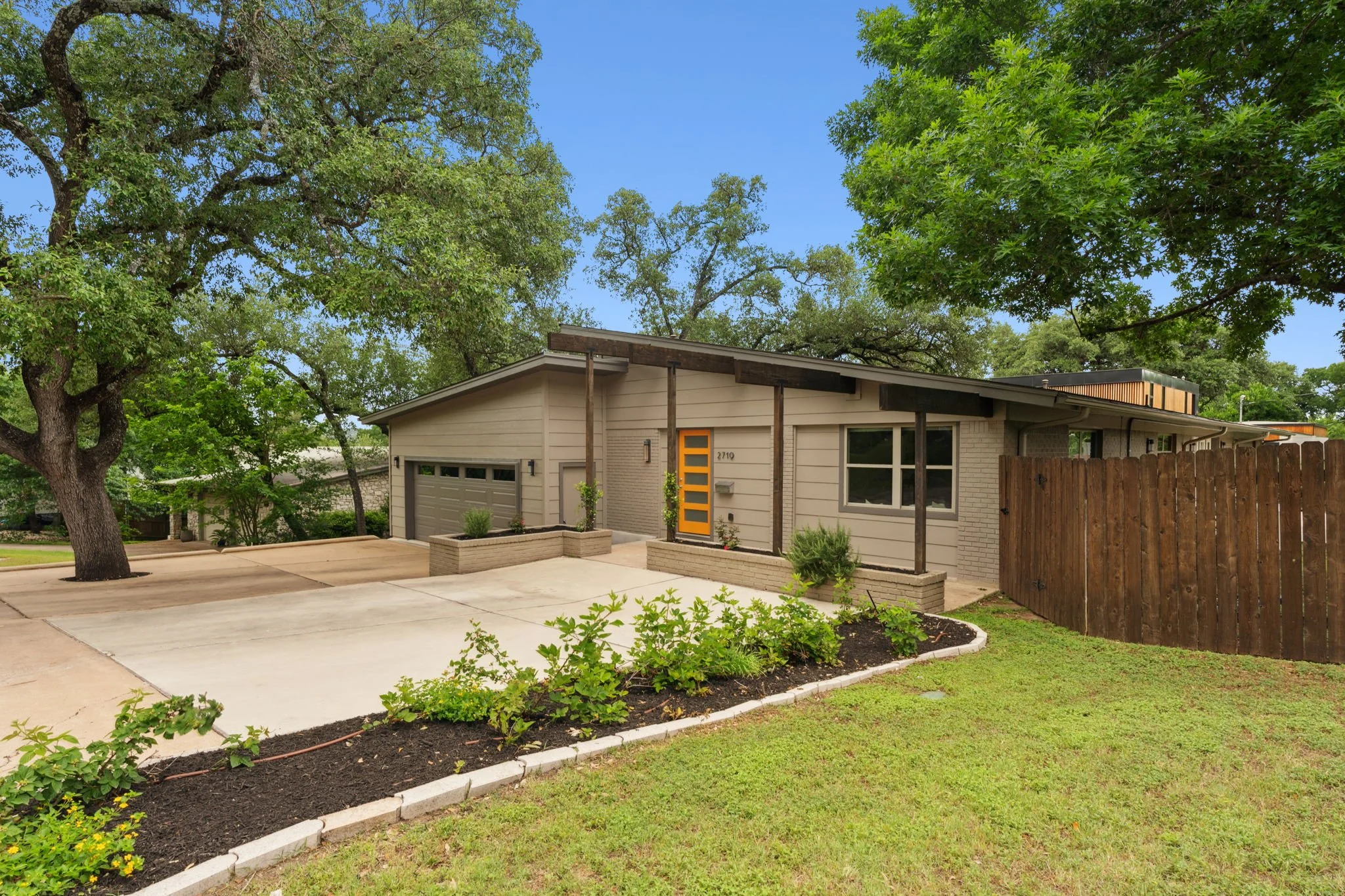 Front view of a modern house with a light-colored exterior, orange door, attached garage, and a landscaped yard with green shrubs and a curved flower bed. Large trees with green leaves surround the house under a clear blue sky.
