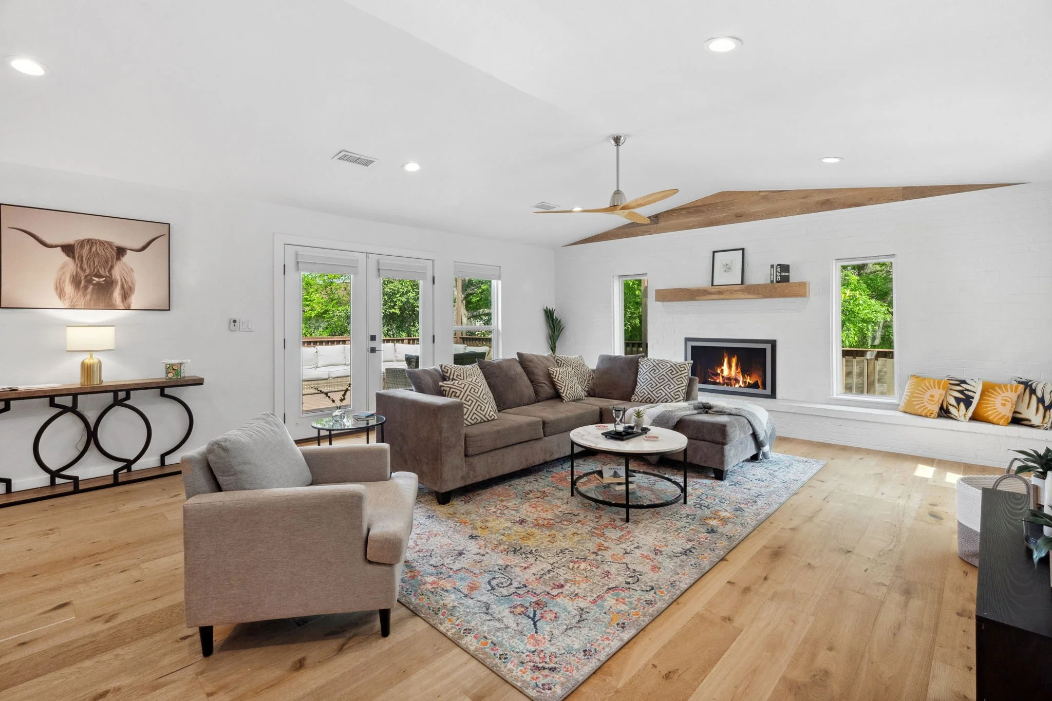 Living room with wood flooring, a gray sectional sofa, armchair, area rug, coffee tables, a fireplace, large windows, art on the wall, a clock, shelves, trees outside, and ceiling lights.