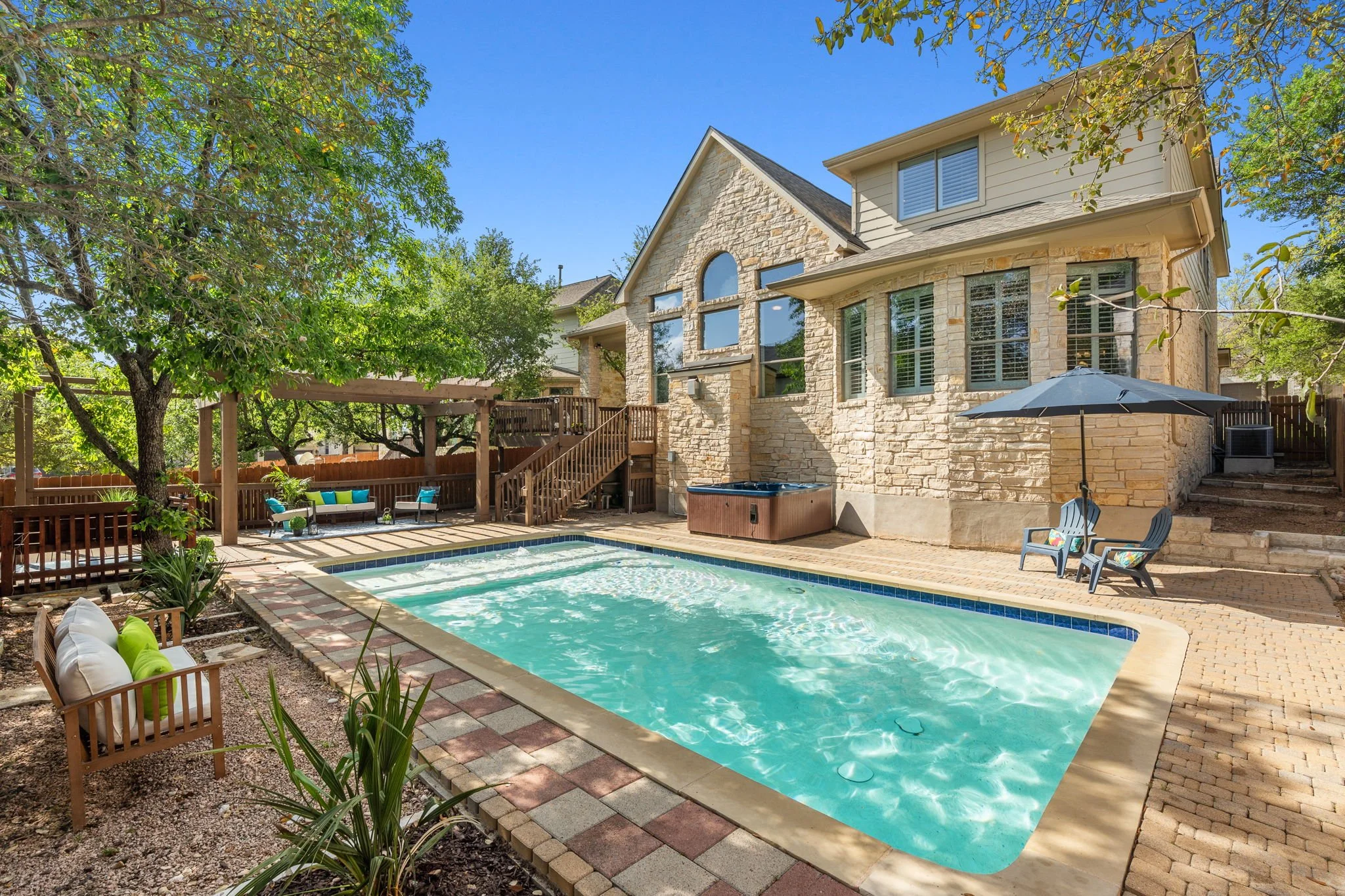 A backyard with a swimming pool, outdoor seating area with cushions, a large tree, and a two-story house with stone and siding exterior and multiple windows, under a clear blue sky.