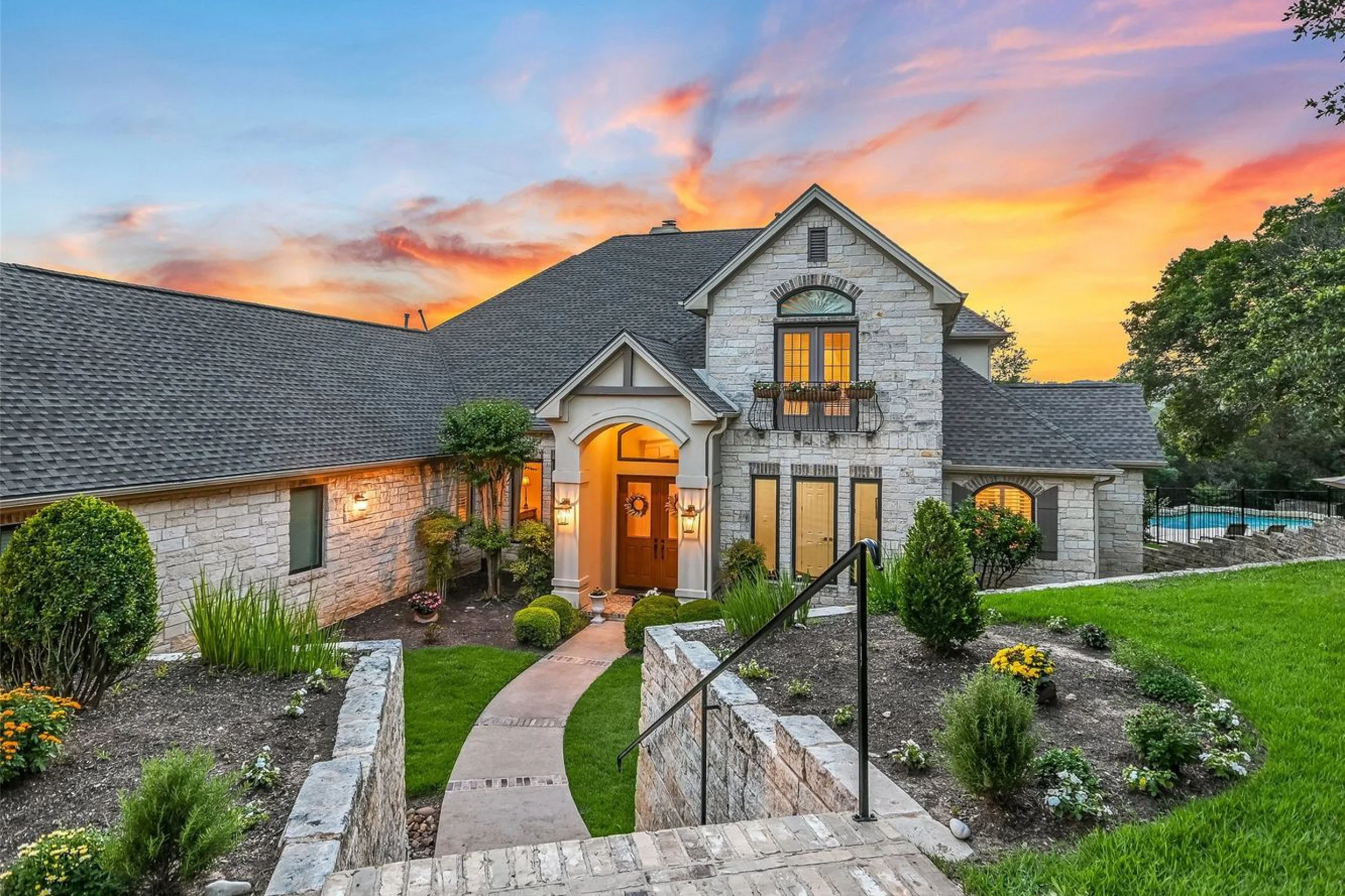 A two-story house with a stone facade, gabled roof, and lit windows at sunset, with a landscaped front yard featuring a curved walkway, green grass, bushes, and flowers.